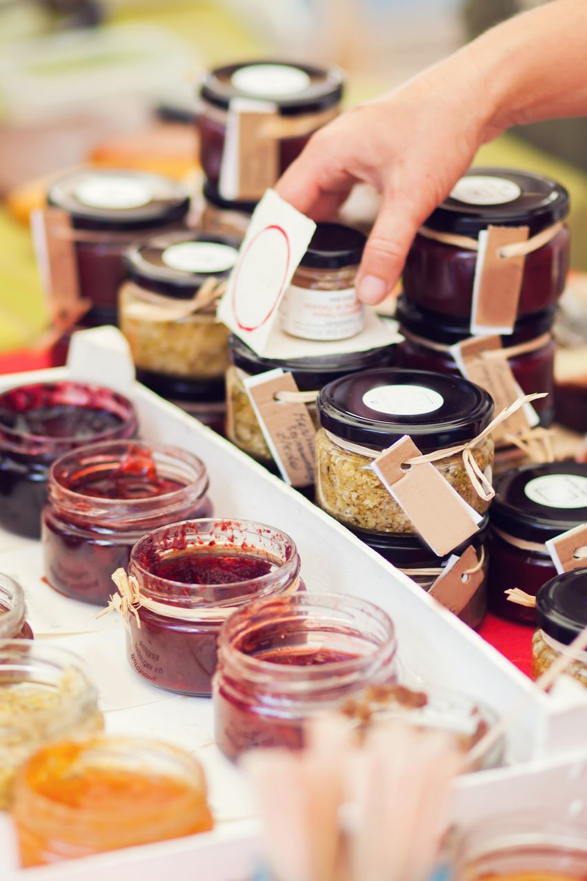 Une main se tend vers un pot de confiture parmi de nombreux autres présentés dans de petits récipients en verre à couvercle sur un plateau blanc.