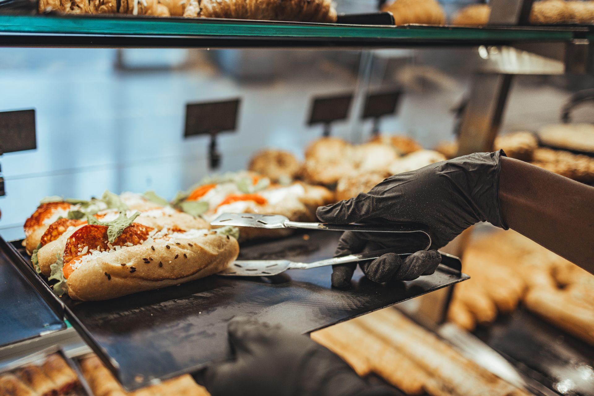Dans une boulangerie, une personne portant des gants noirs utilise des pinces argentées pour disposer un sandwich sur un plateau de présentation.