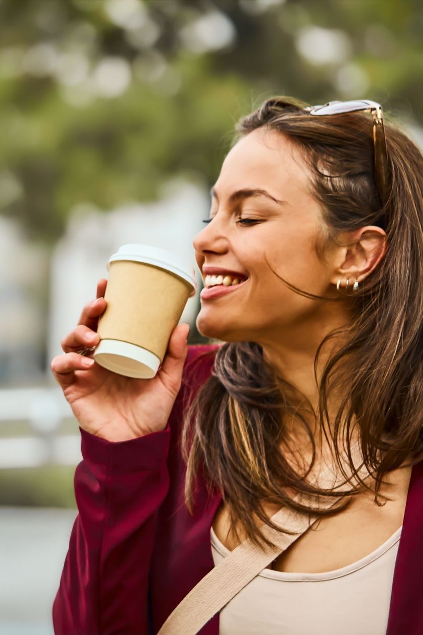 Une personne vêtue d'un cardigan bordeaux sourit en humant l'odeur d'une boisson chaude dans un gobelet en carton beige.
