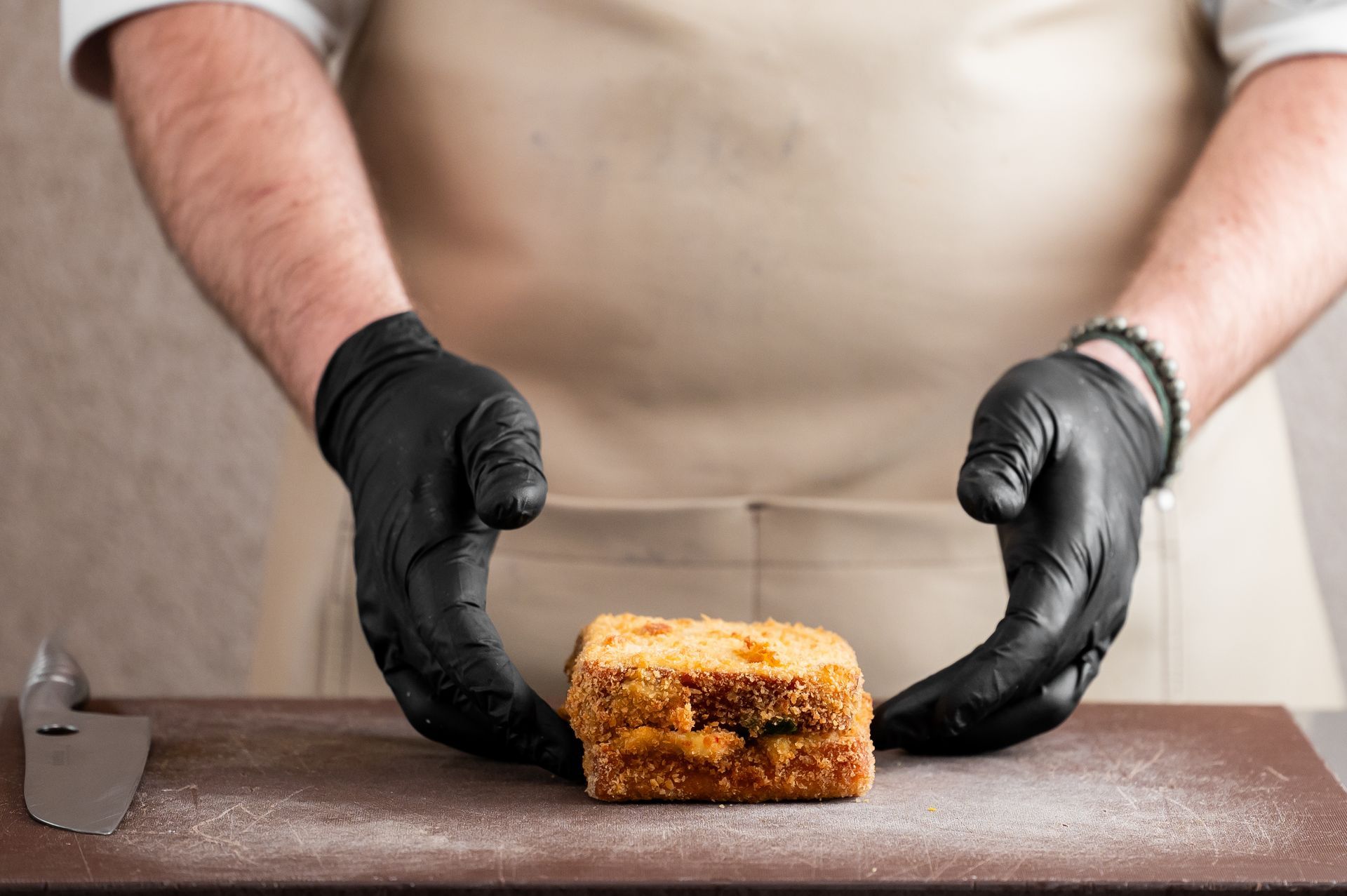 Une personne portant des gants noirs tient délicatement un carré de pâtisserie dorée sur une planche en bois.