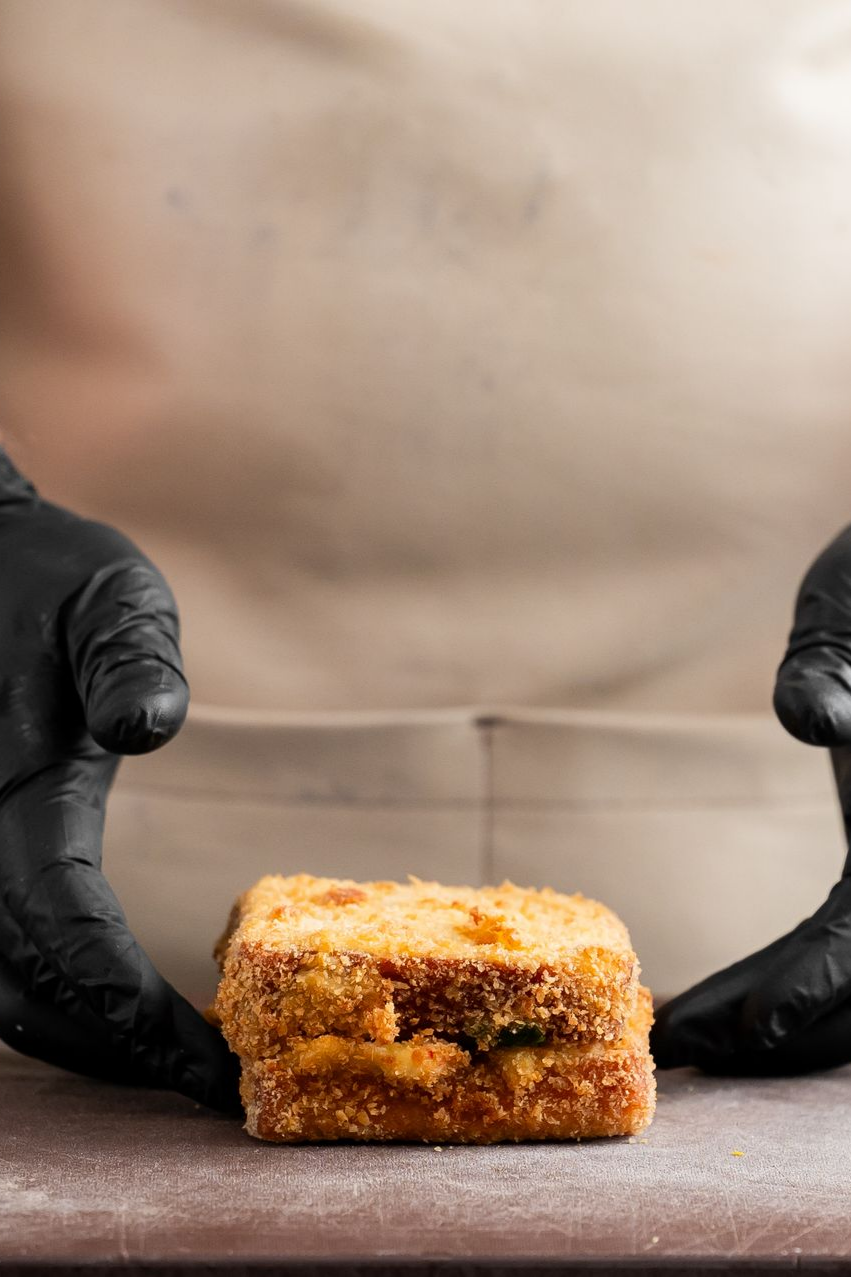 Des mains gantées de noir encadrent un sandwich frit carré et doré, posé sur un plan de travail devant un fond uni.