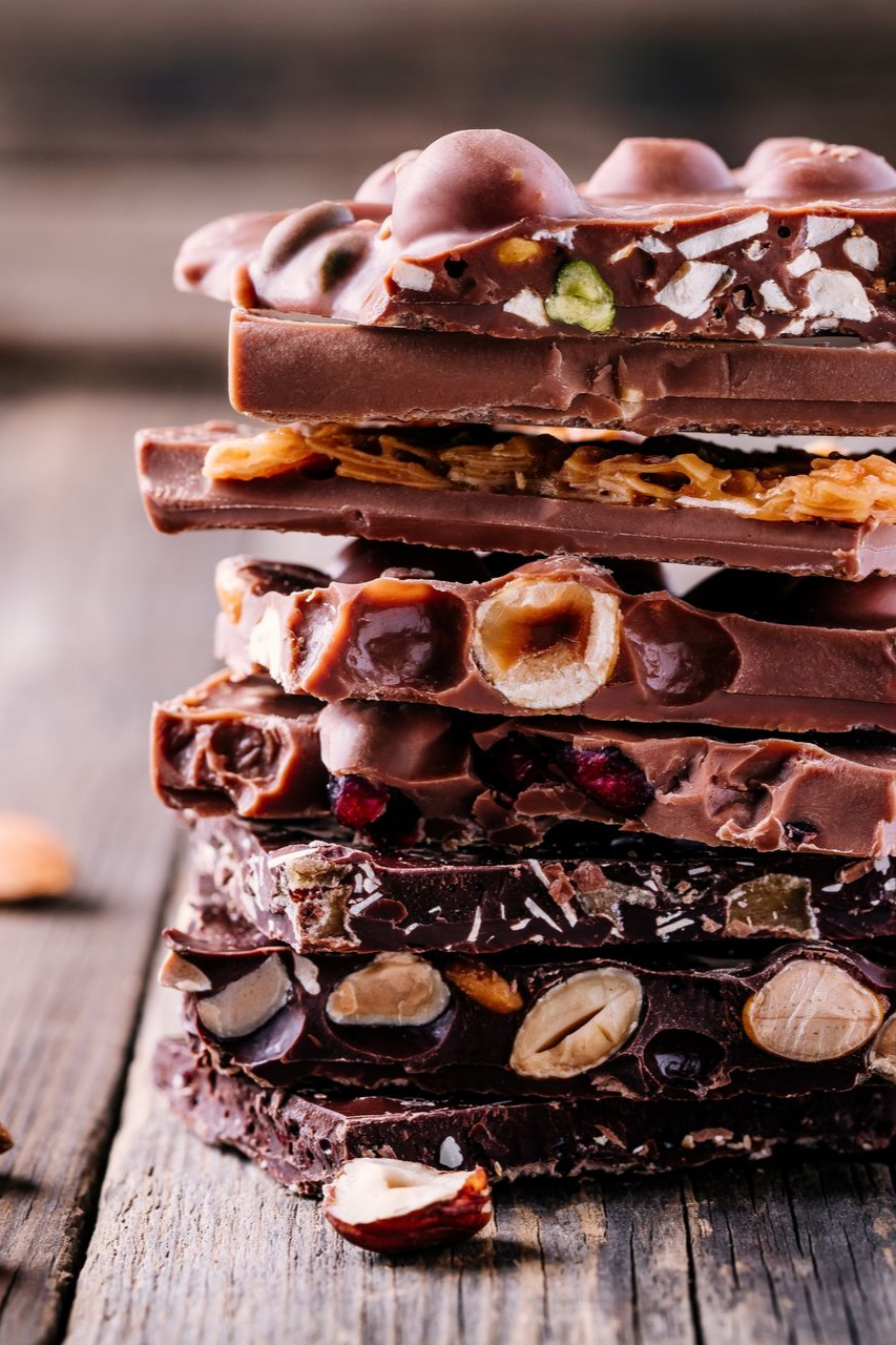 Une pile verticale de barres de chocolat gourmandes variées, agrémentées de noix, de fruits secs et de caramel, posée sur une planche en bois rustique.