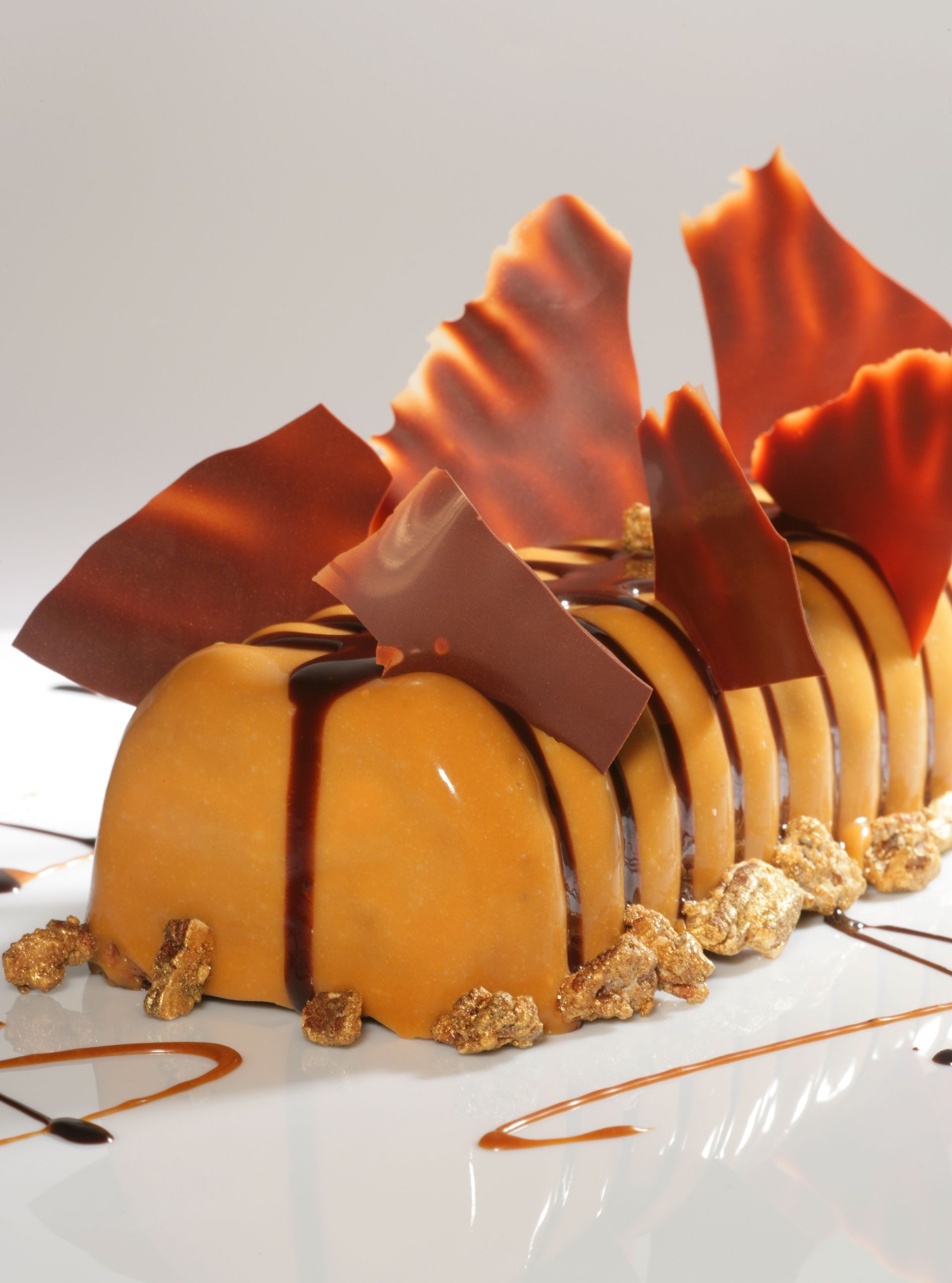 Un dessert rectangulaire glacé au caramel, garni d'éclats de chocolat et de miettes saupoudrées de poudre d'or, présenté sur une assiette blanche.