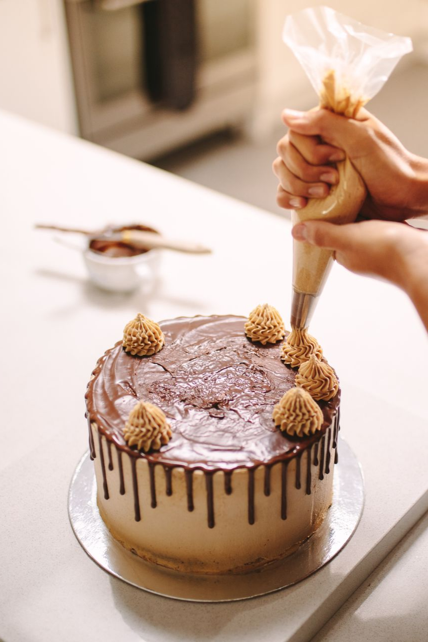 Des mains utilisent une poche à douille pour ajouter des volutes décoratives de crème au beurre sur le dessus d'un gâteau au chocolat nappé, posé sur un comptoir de cuisine.