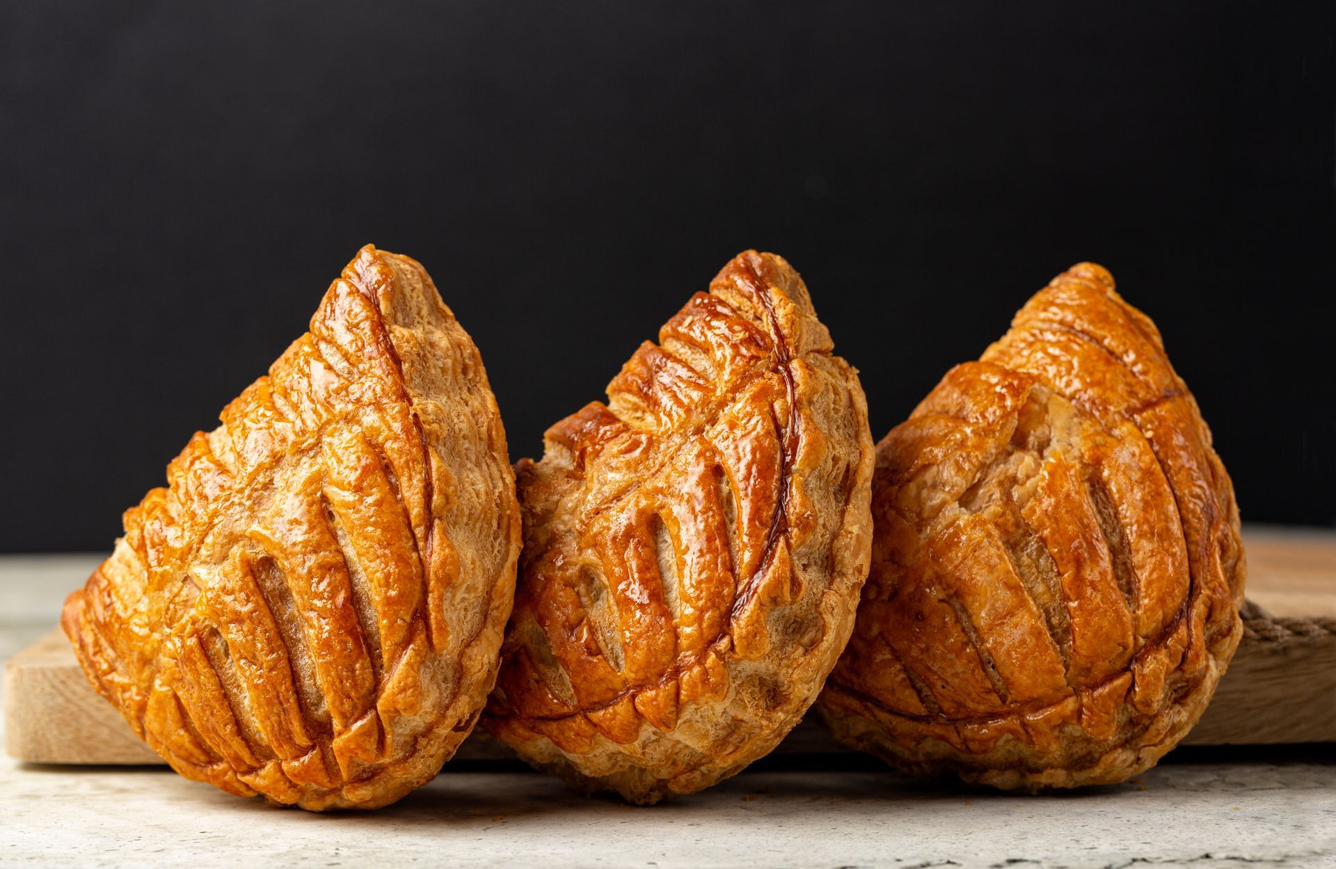 Trois chaussons aux pommes dorés, décorés de motifs en forme de feuilles, sont alignés sur une planche en bois, sur fond sombre.