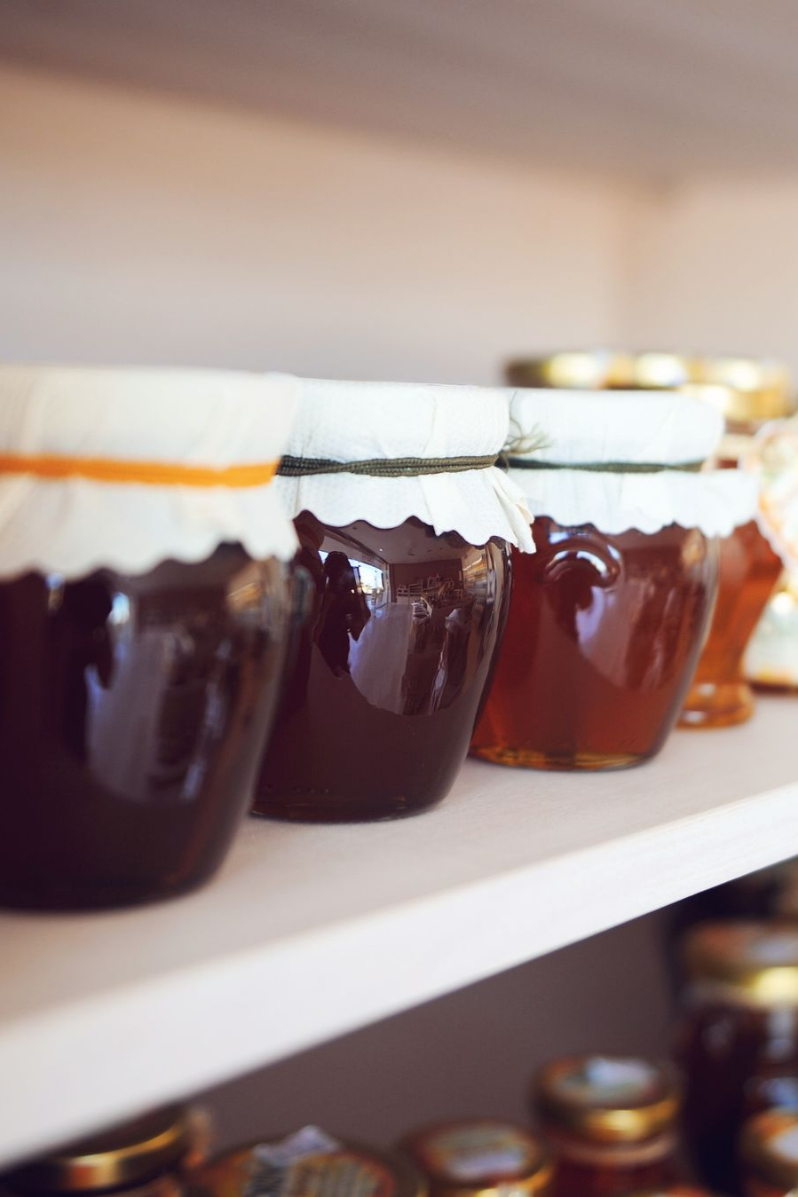Des pots en verre contenant du miel ambré foncé, recouverts de tissu blanc, sont disposés sur des étagères en bois blanc.