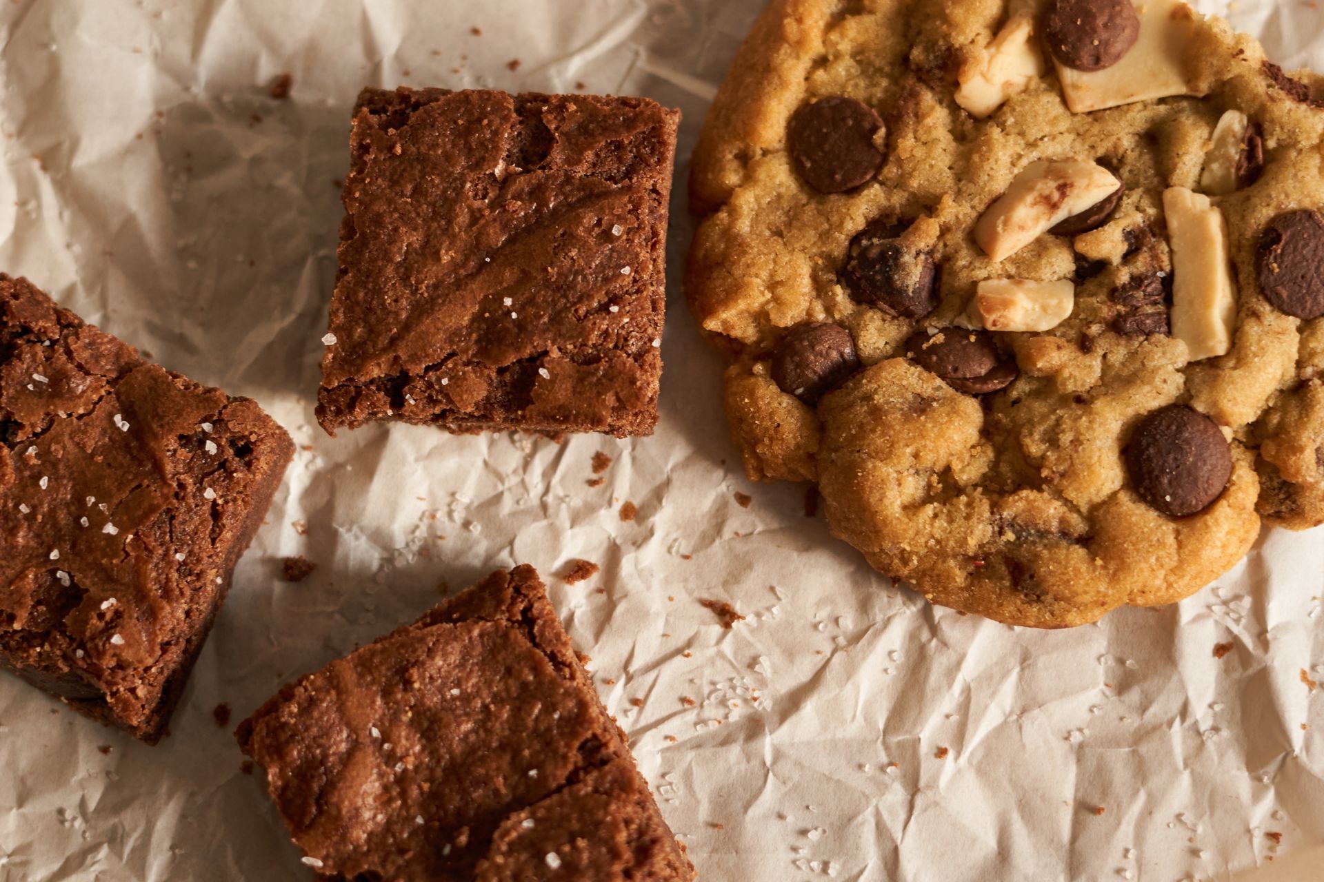 Trois brownies carrés au chocolat et un grand cookie aux pépites de chocolat disposés sur du papier sulfurisé blanc froissé.