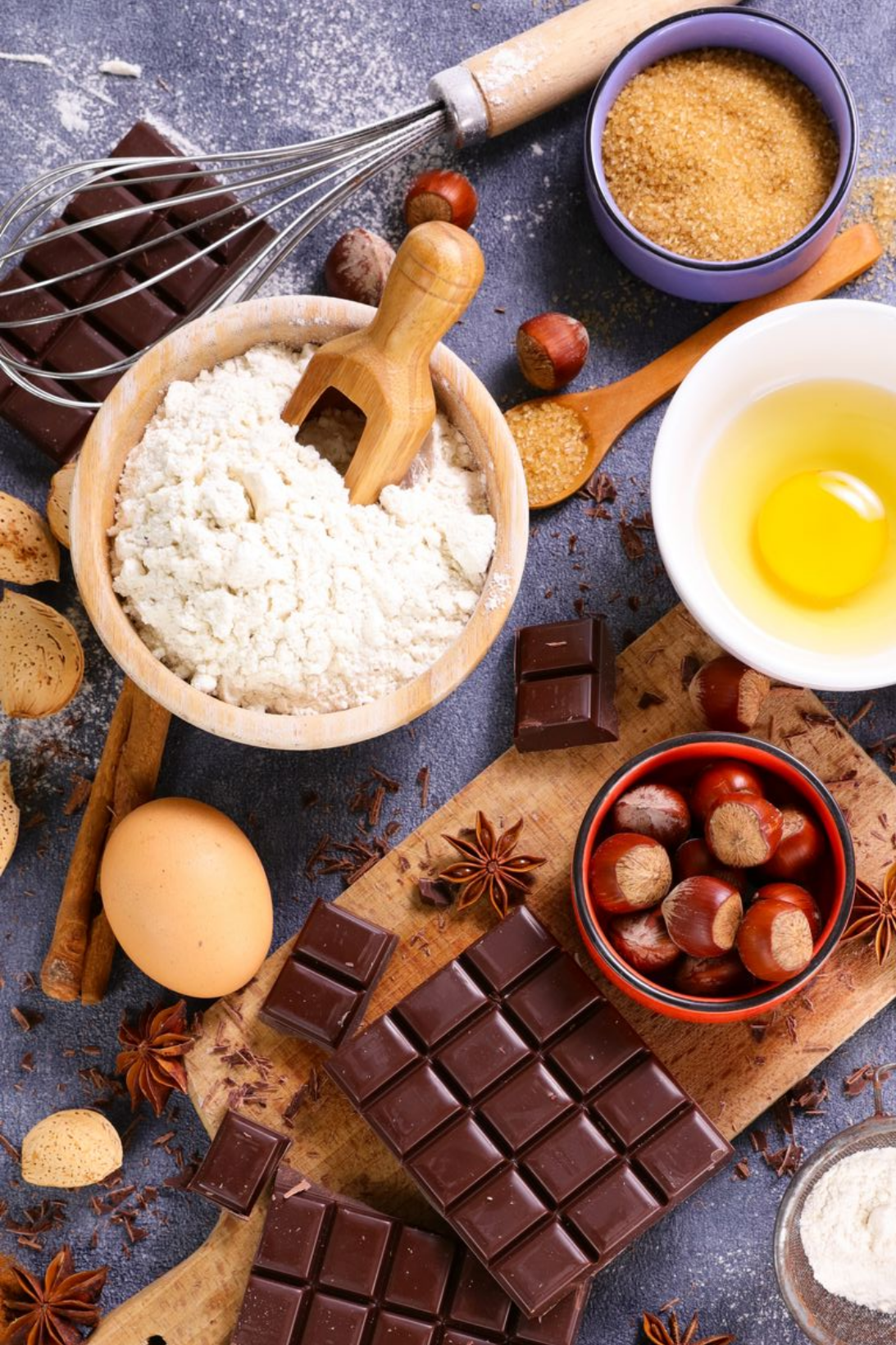 Ingrédients pour la pâtisserie, notamment chocolat noir, farine, œufs crus, noisettes et épices, disposés sur une planche en bois rustique.
