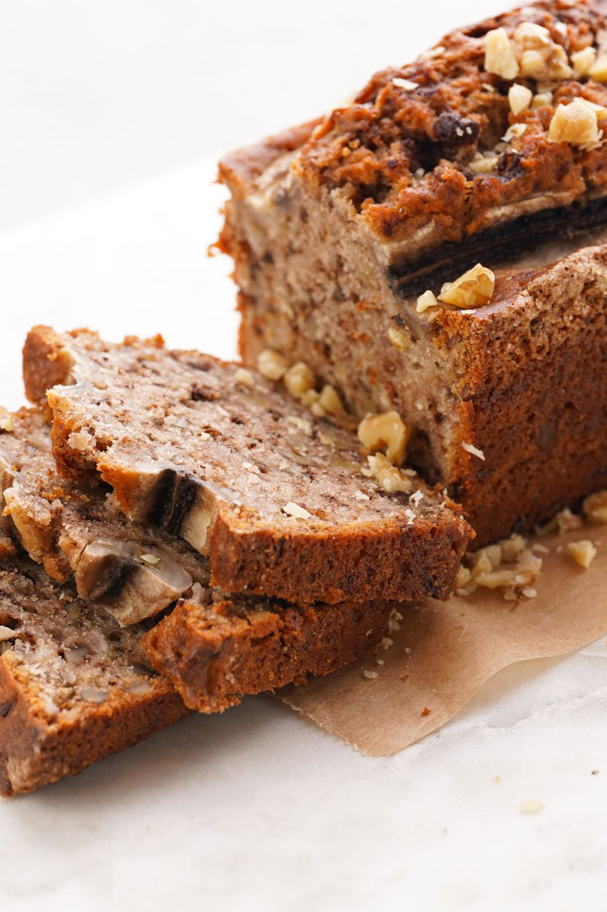 Un cake aux bananes et aux noix, coupé en deux tranches épaisses, servi sur du papier kraft devant une surface en marbre blanc.