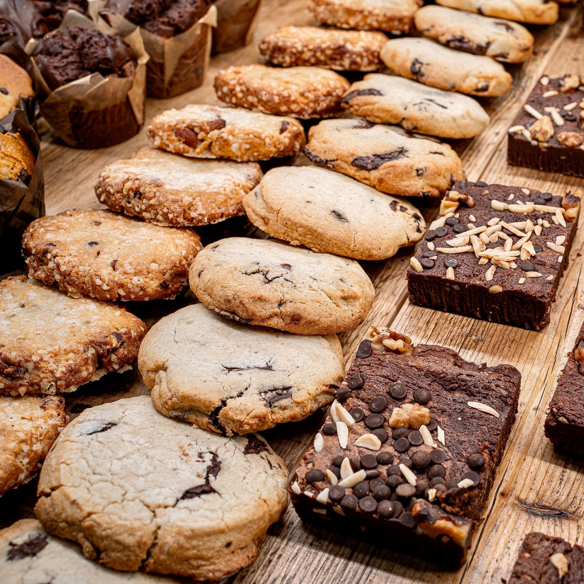 Un assortiment de cookies aux pépites de chocolat, de brownies garnis de noix et de pépites, et de muffins présentés sur une surface en bois.