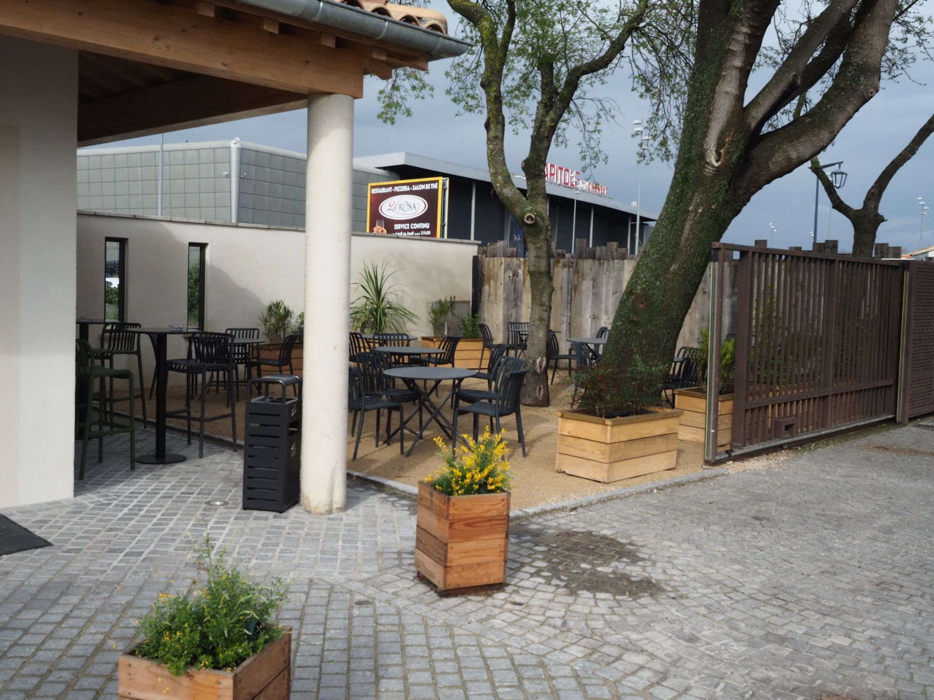 Une terrasse extérieure avec des tables et des chaises de café, des jardinières en bois et des arbres, jouxtant un bâtiment couvert.