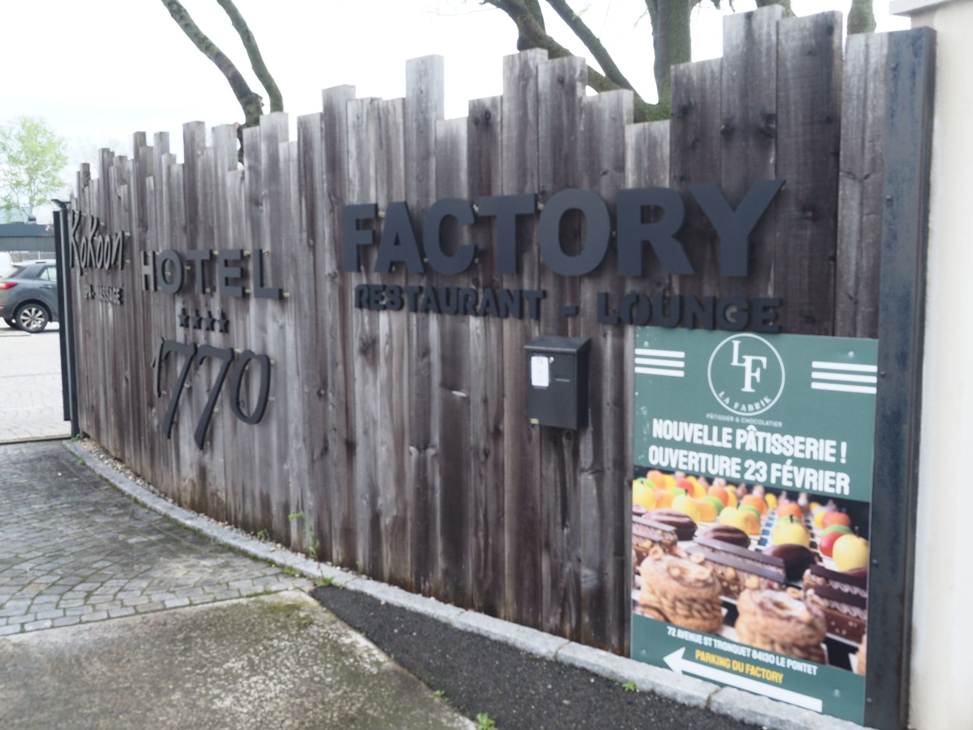Une clôture en bois patiné affiche en lettres noires les inscriptions Factory Restaurant Lounge et Hotel 770 au-dessus d'une enseigne de pâtisserie.