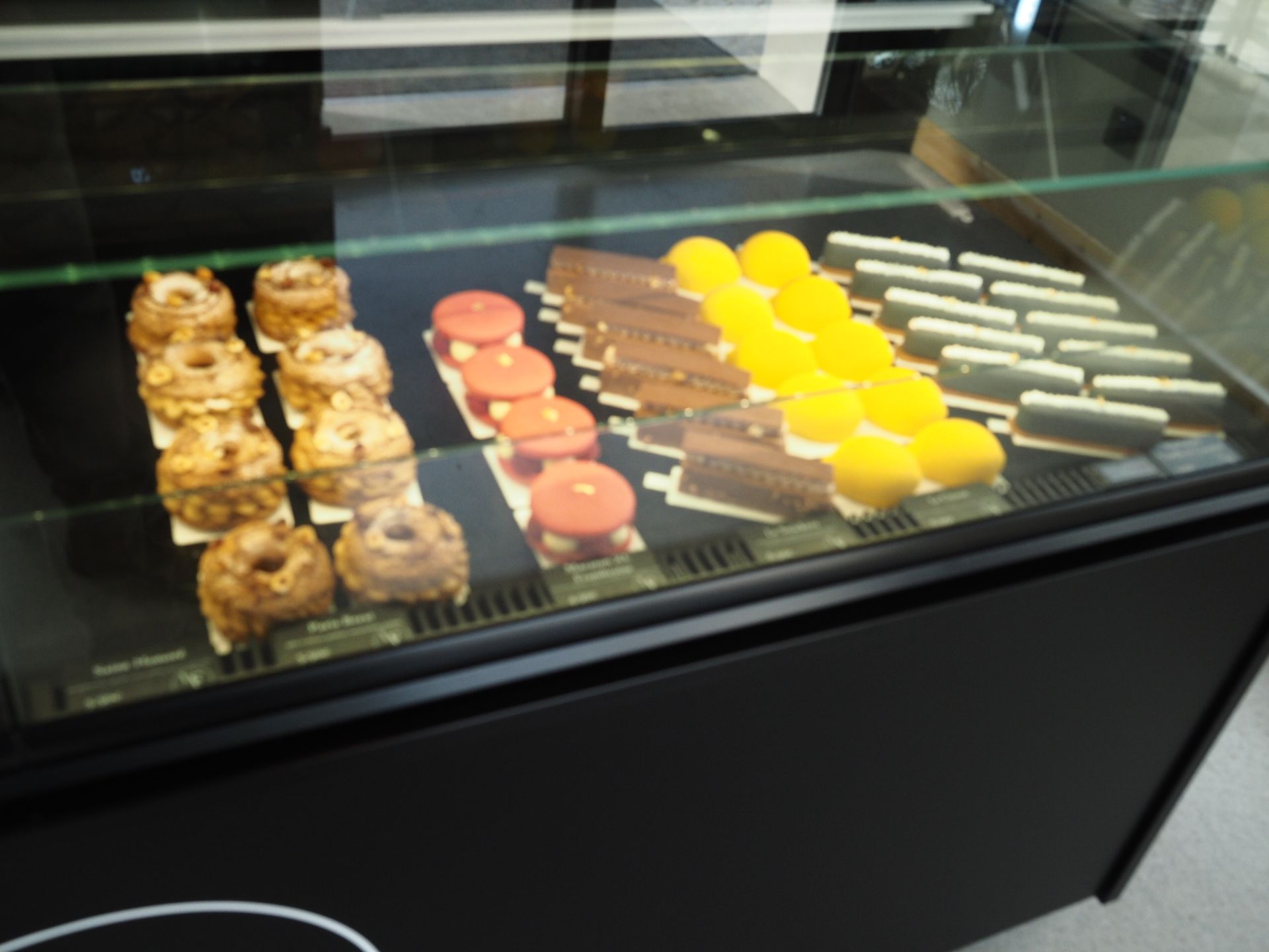 Une vitrine présentant des rangées de pâtisseries variées, notamment des beignets glacés, des macarons roses, des dômes jaunes et des brownies.