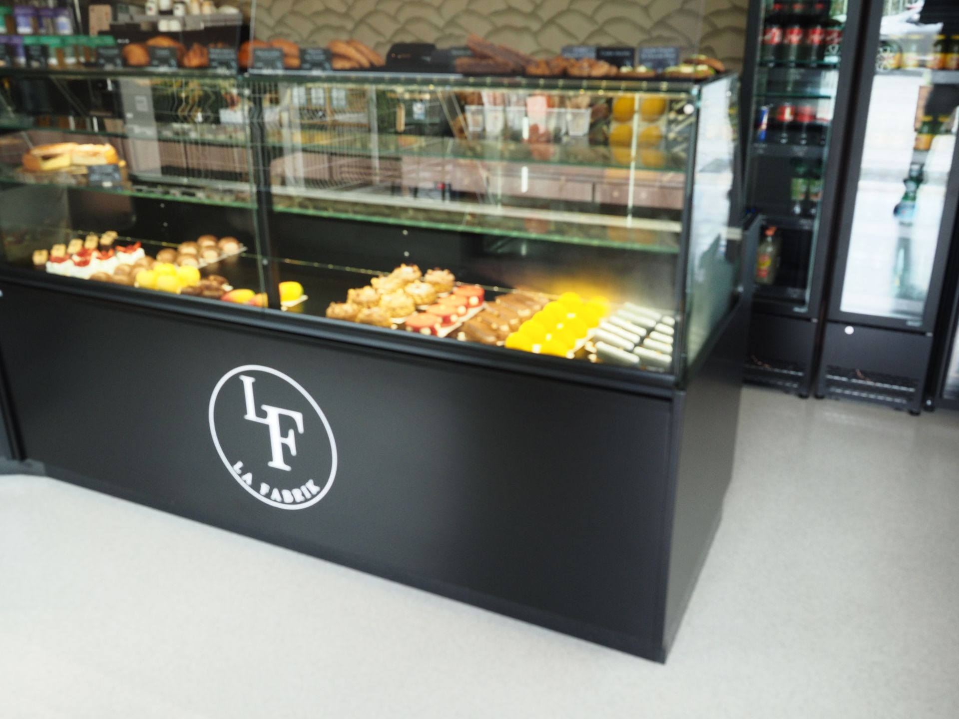 Une vitrine en verre dans une boulangerie, remplie de pâtisseries et de desserts variés, avec un socle noir et un logo blanc.