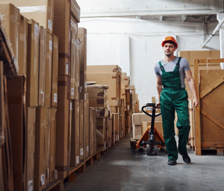 Ein Arbeiter in einem grünen Overall und einem orangefarbenen Schutzhelm geht durch ein Lagerhaus und zieht einen mit Kisten beladenen Hubwagen hinter sich her.