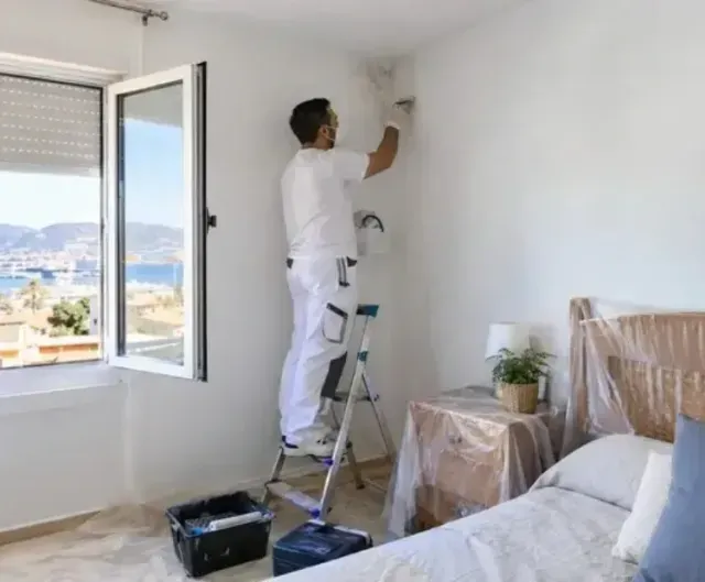Pintor subido a una escalera pintando la pared blanca de un dormitorio junto a una ventana con vistas a la ciudad