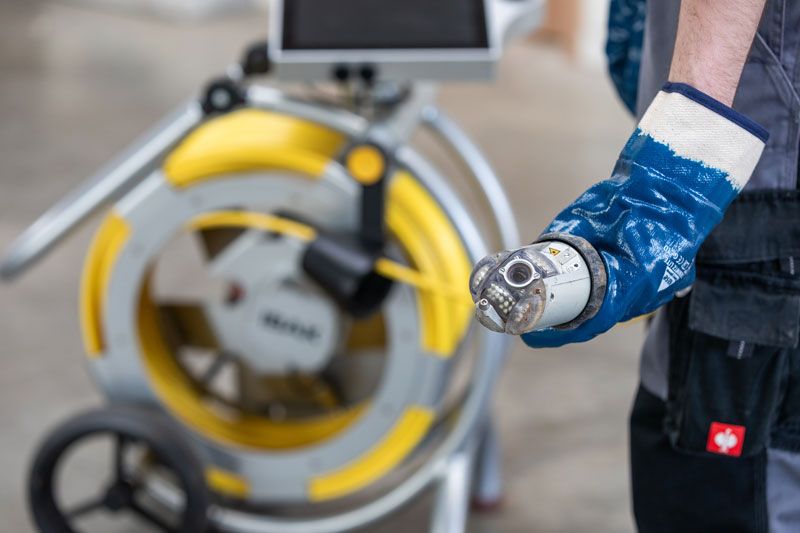Ein Klempner benutzt eine Abflussreinigungsmaschine auf dem Küchenboden. Rote Maschine, schwarzer Schlauch, Metallgitter.