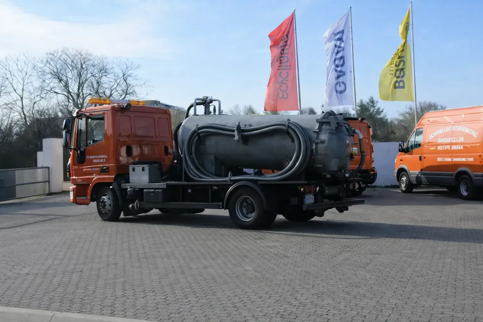 Ein orangefarbener Abwasserwagen mit großem Tank steht auf einer gepflasterten Fläche; im Hintergrund sind Flaggen zu sehen.