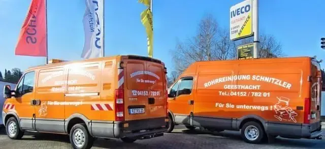 Zwei orangefarbene Lieferwagen mit Aufschrift. Im Hintergrund ein IVECO-Schild und Flaggen. Blauer Himmel.