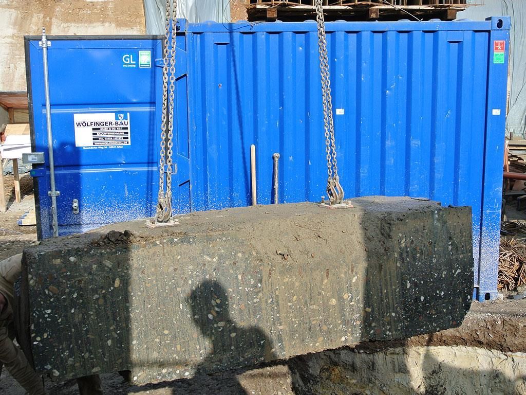 Ein großer Betonblock wird an Ketten vor einen blauen Container gehoben