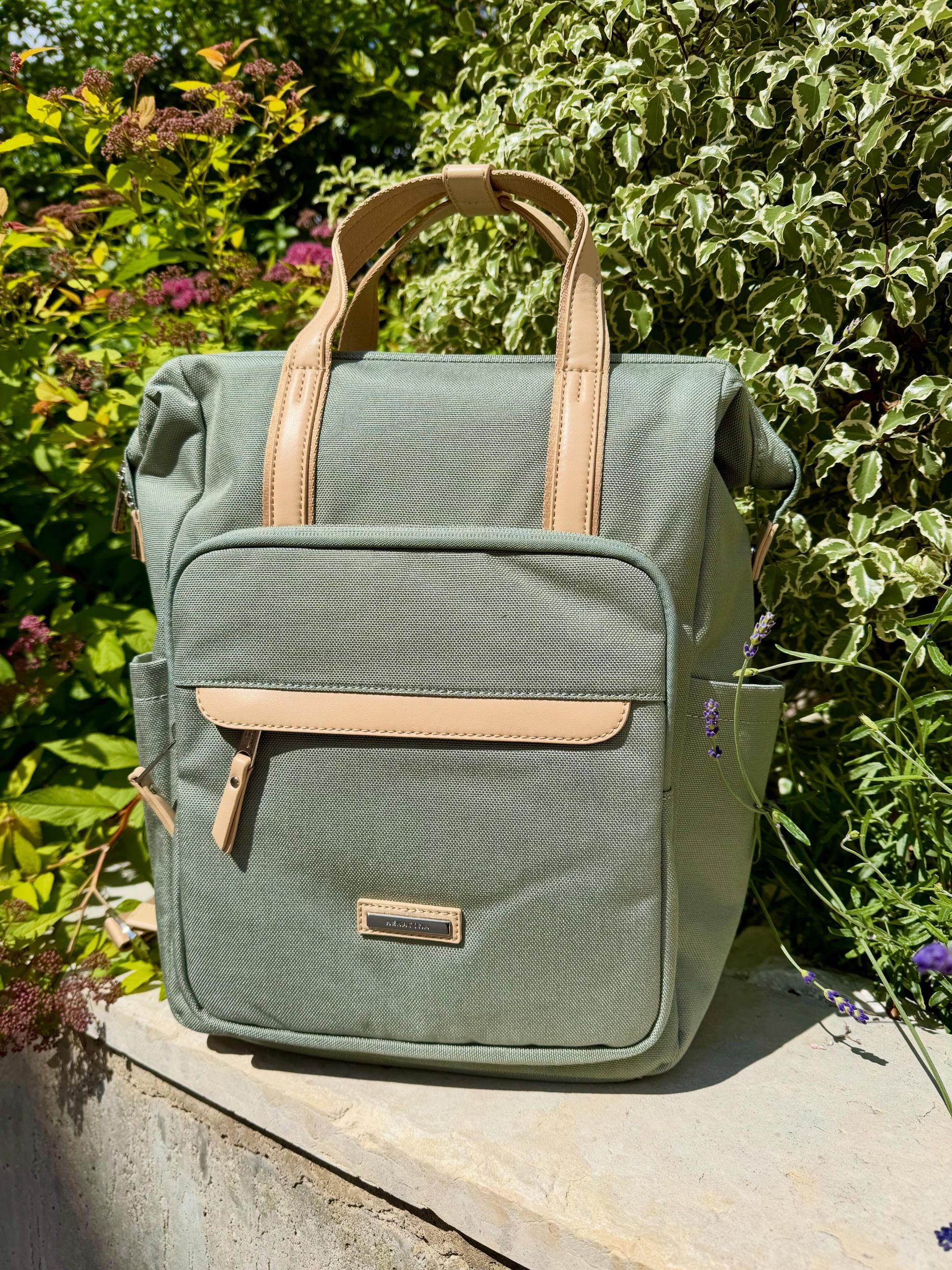 Un sac à dos matelassé vert à bretelles beiges est posé à l'extérieur, sur un rebord de pierre, avec des fleurs en arrière-plan.
