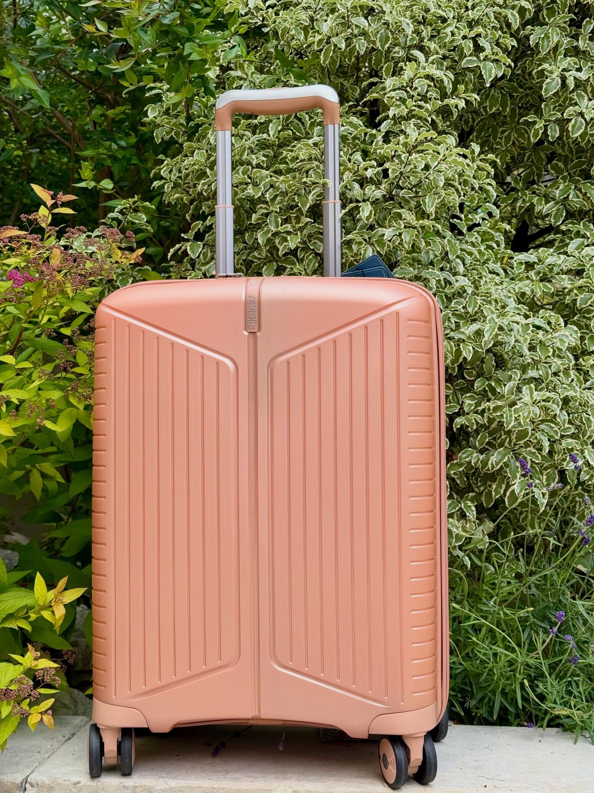 Valise cabine rigide couleur pêche, posée debout sur un chemin extérieur bordé d'arbres.