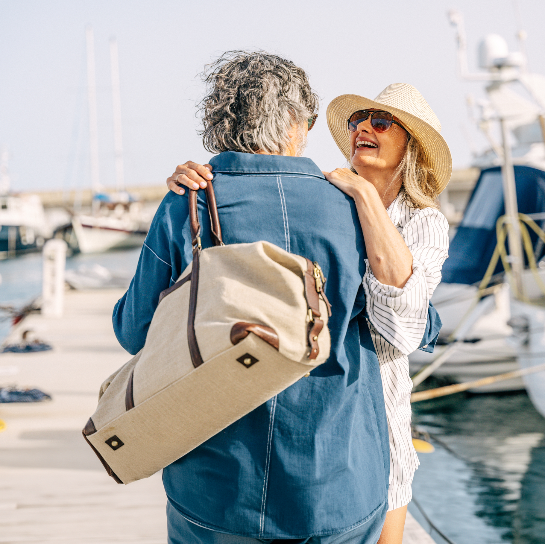 Deux personnes sourient près d'un port de plaisance, l'une avec un sac à dos beige et un chapeau de paille, des bateaux en arrière-plan.