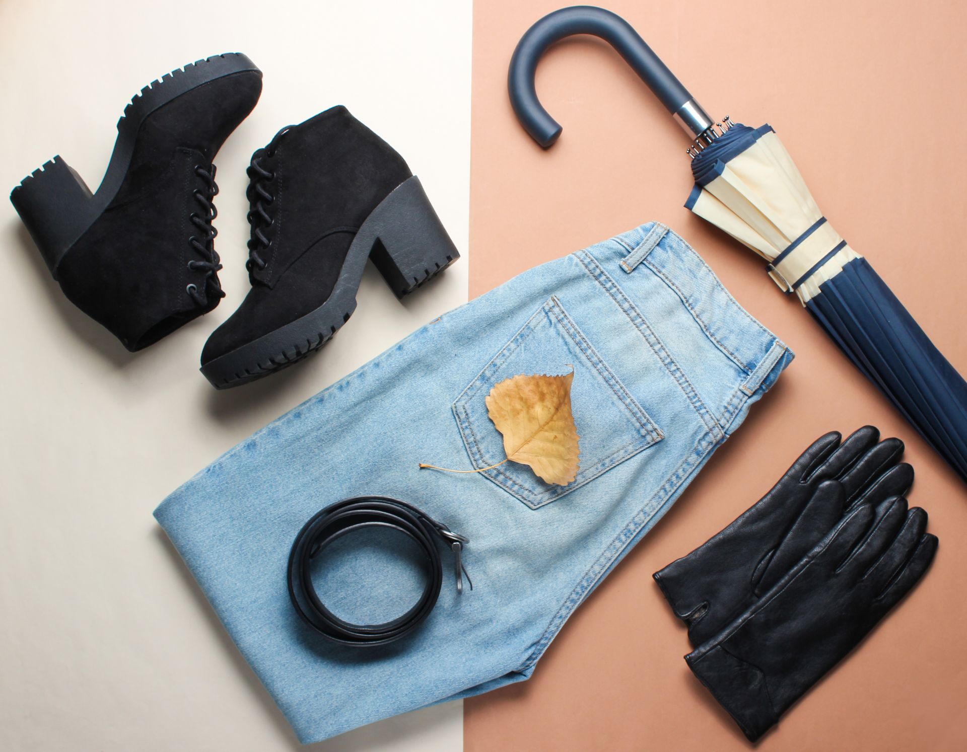 Chaussures noires, gants noirs, jean bleu et parapluie plié disposés sur un fond beige.