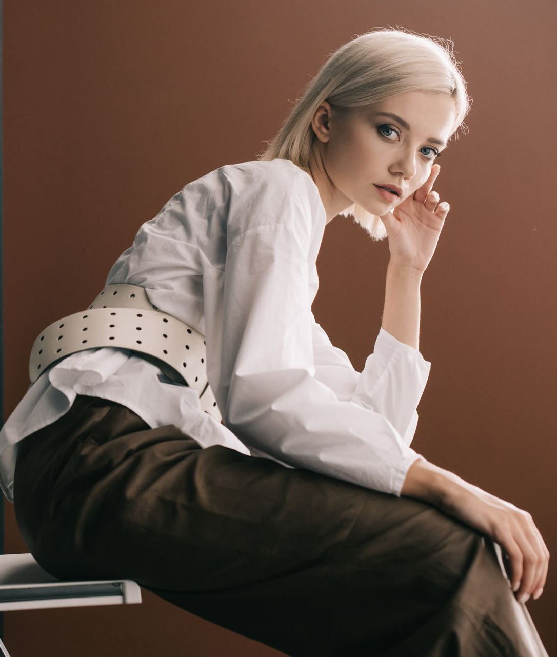 Personne en chemise blanche et pantalon foncé, posant assise devant un fond marron.