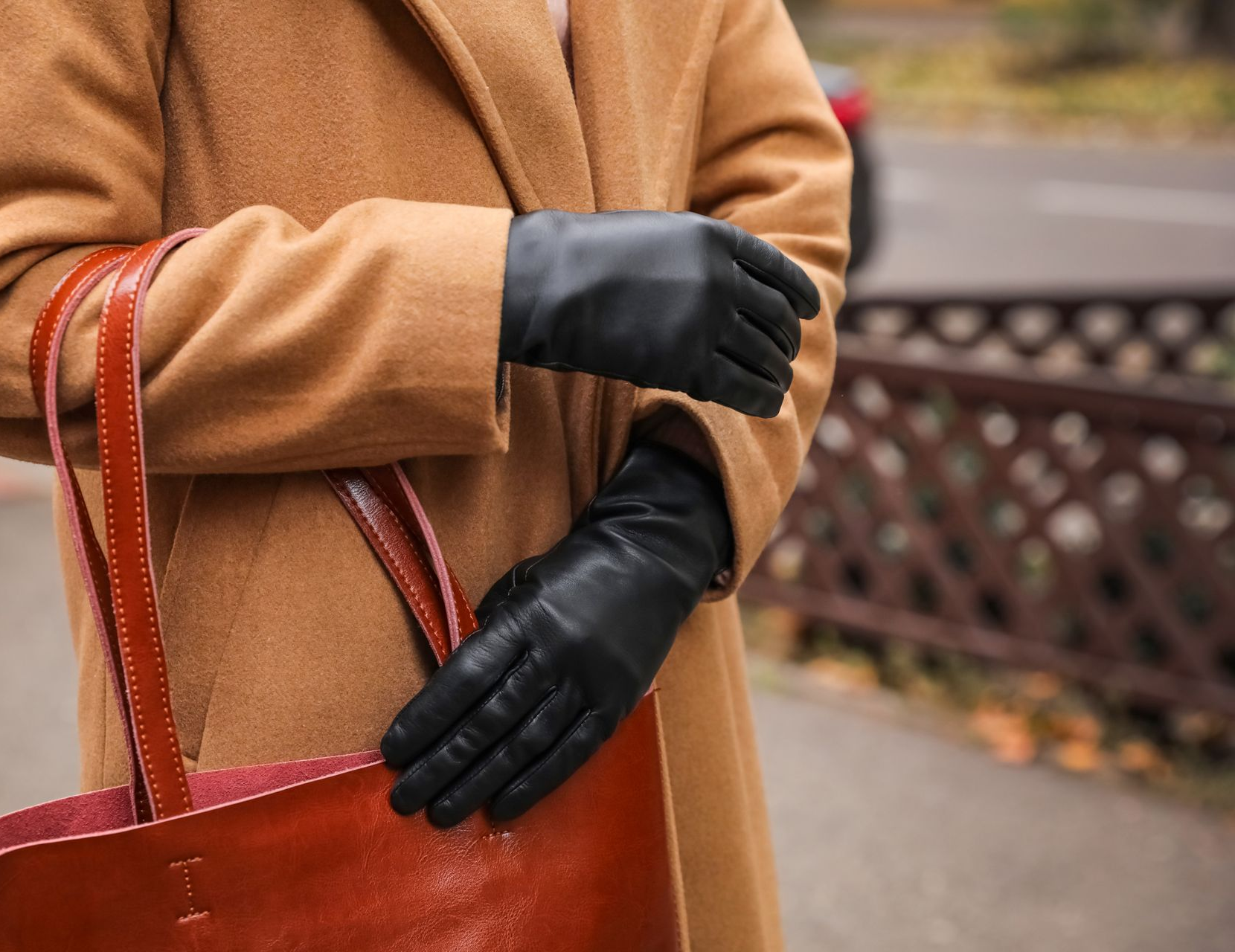 Une personne en manteau beige ajuste des gants noirs à côté d'un sac en cuir rouge à l'extérieur.