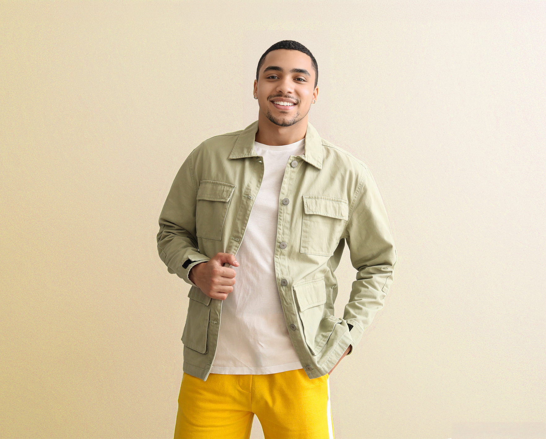 Un homme se tient debout contre un mur beige, portant une veste vert clair, une chemise blanche et un pantalon jaune vif.