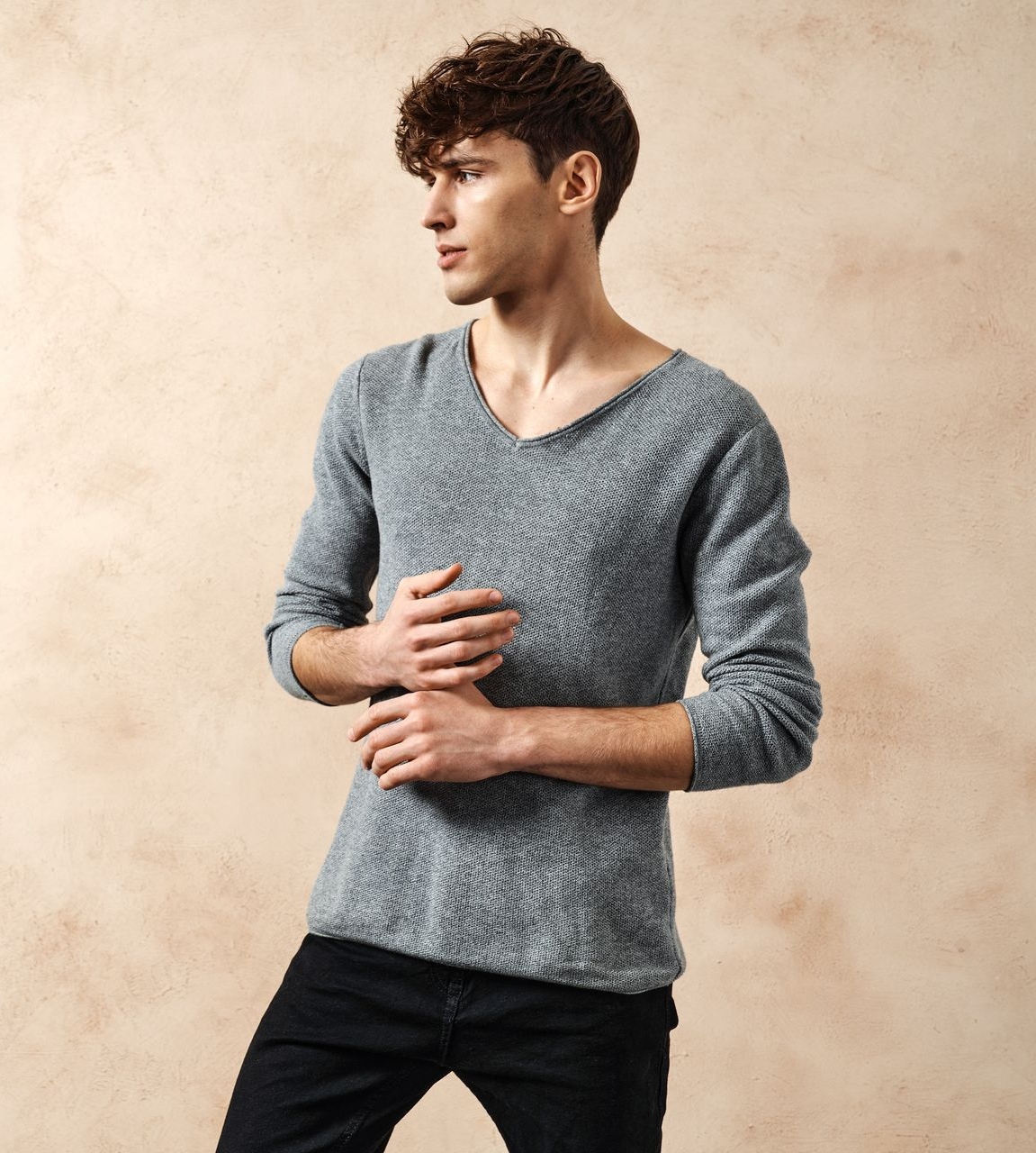 Jeune personne en pull gris se tenant sur un fond beige, regardant sur le côté.
