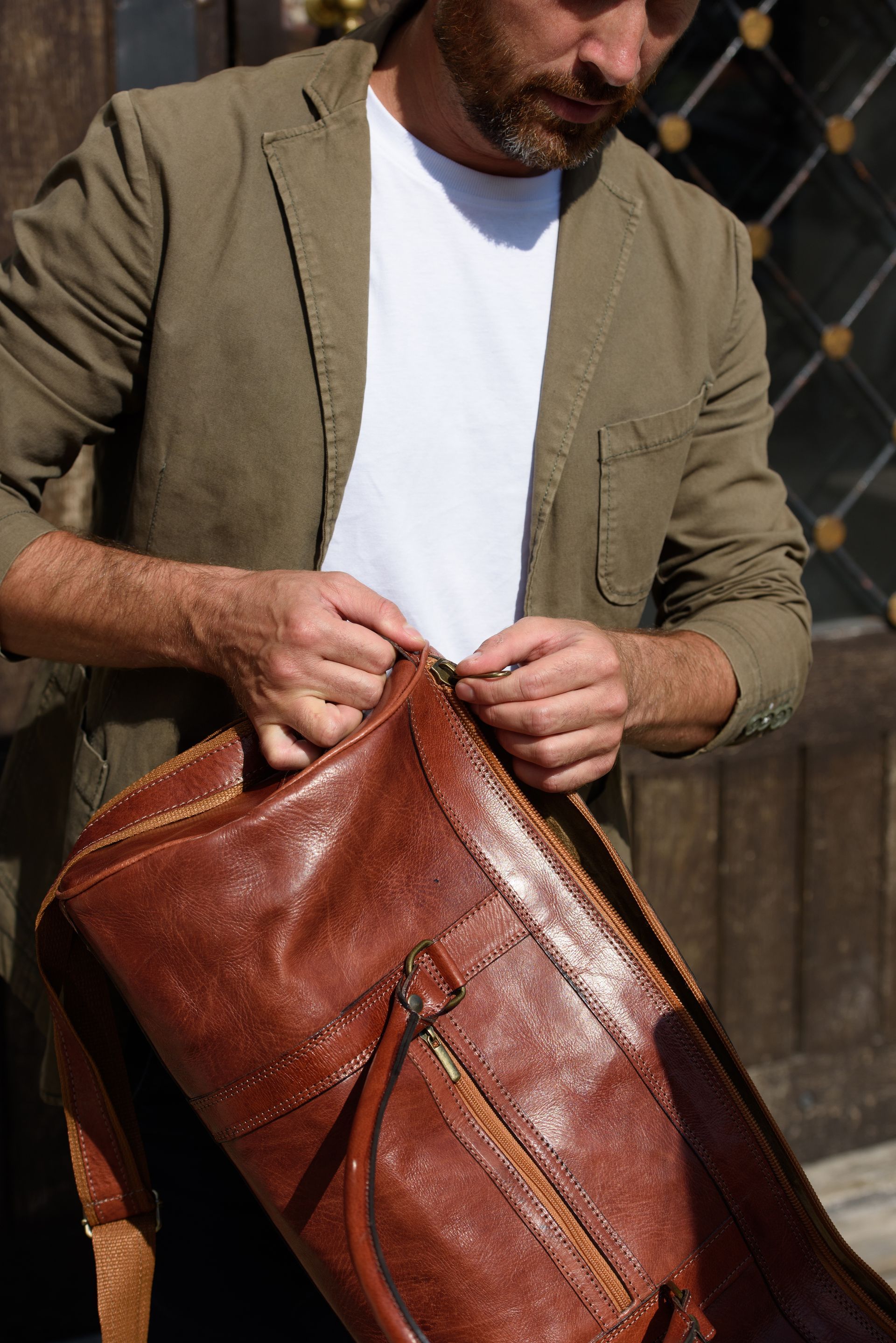 Un homme ajuste un sac en cuir marron contre une clôture en bois à l'extérieur.
