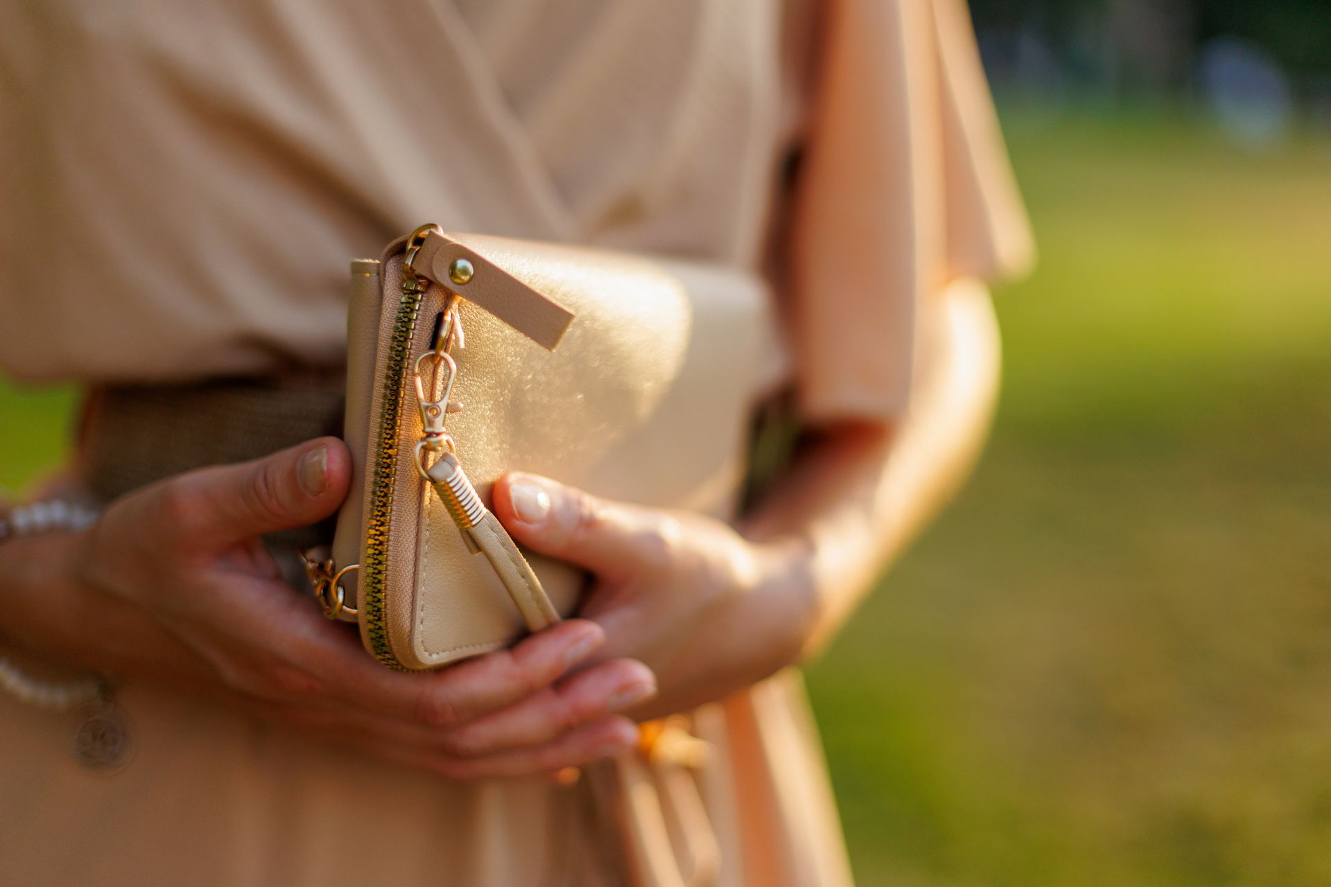 Une personne portant une robe beige et tenant une pochette dorée à l'extérieur, sous une douce lumière du soleil.