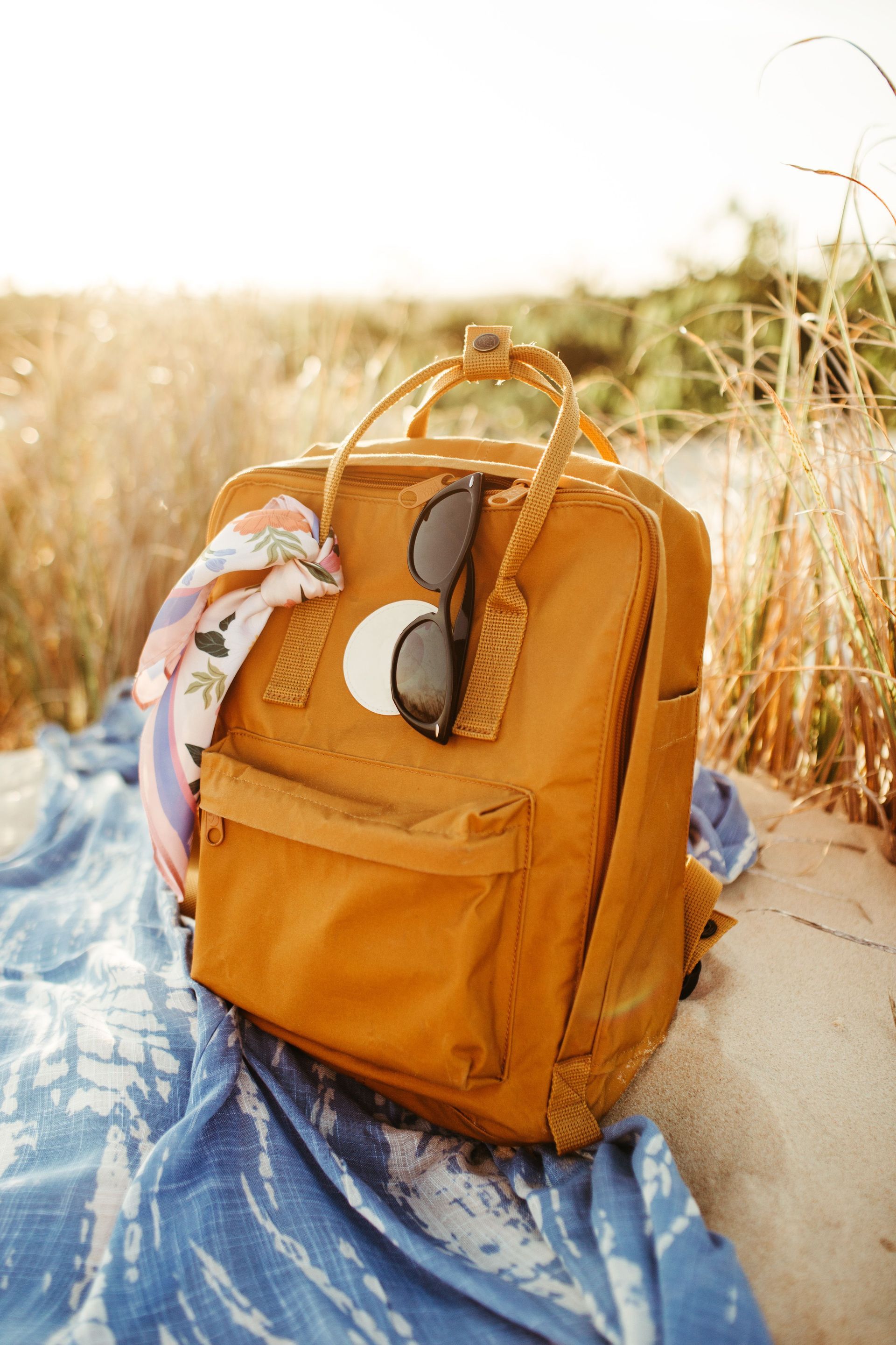 Un sac à dos orange posé sur une couverture bleue dans les hautes herbes, avec une écharpe drapée à côté.