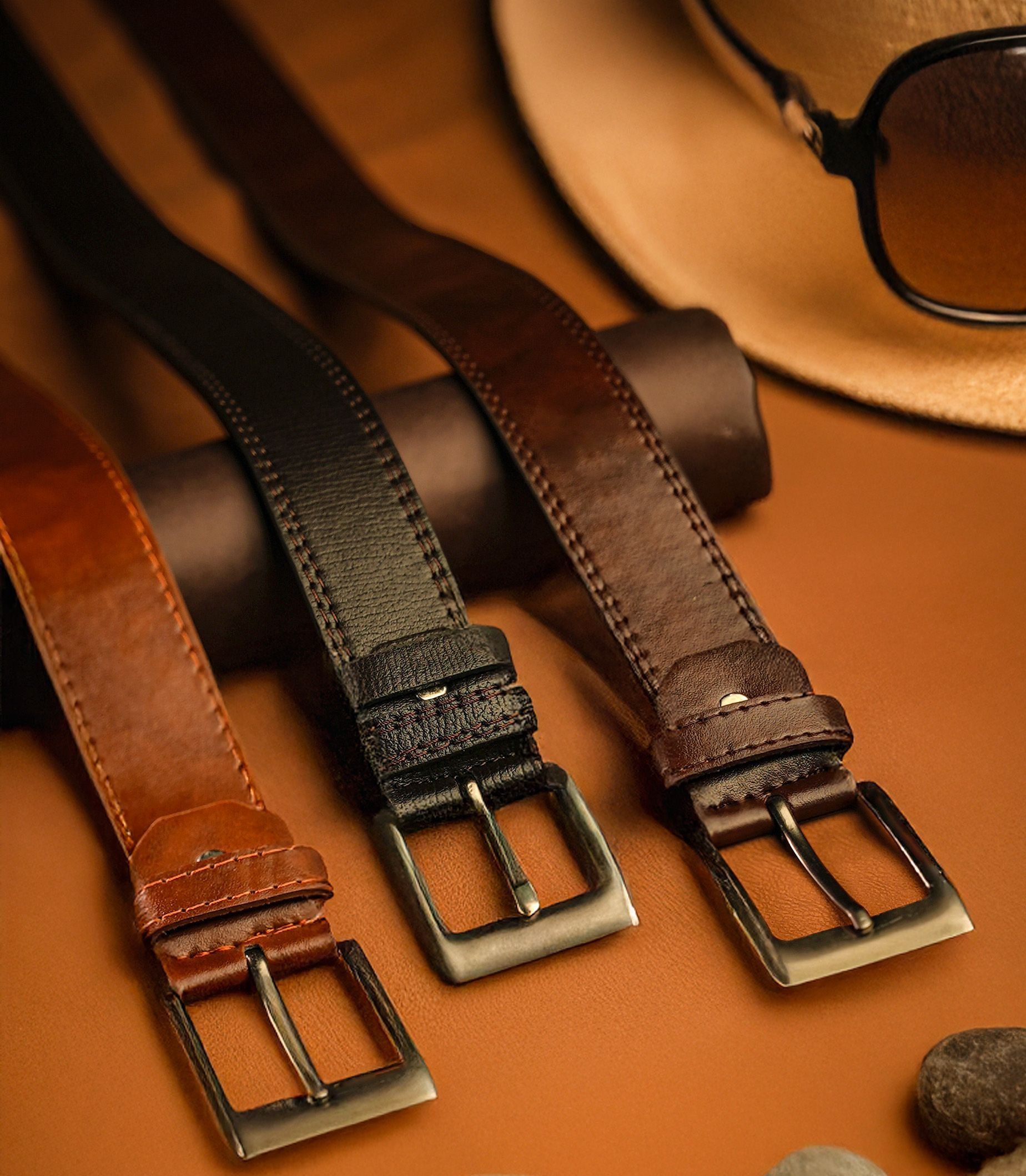 Trois ceintures en cuir avec boucles métalliques marron, noir et marron foncé sur une surface beige.