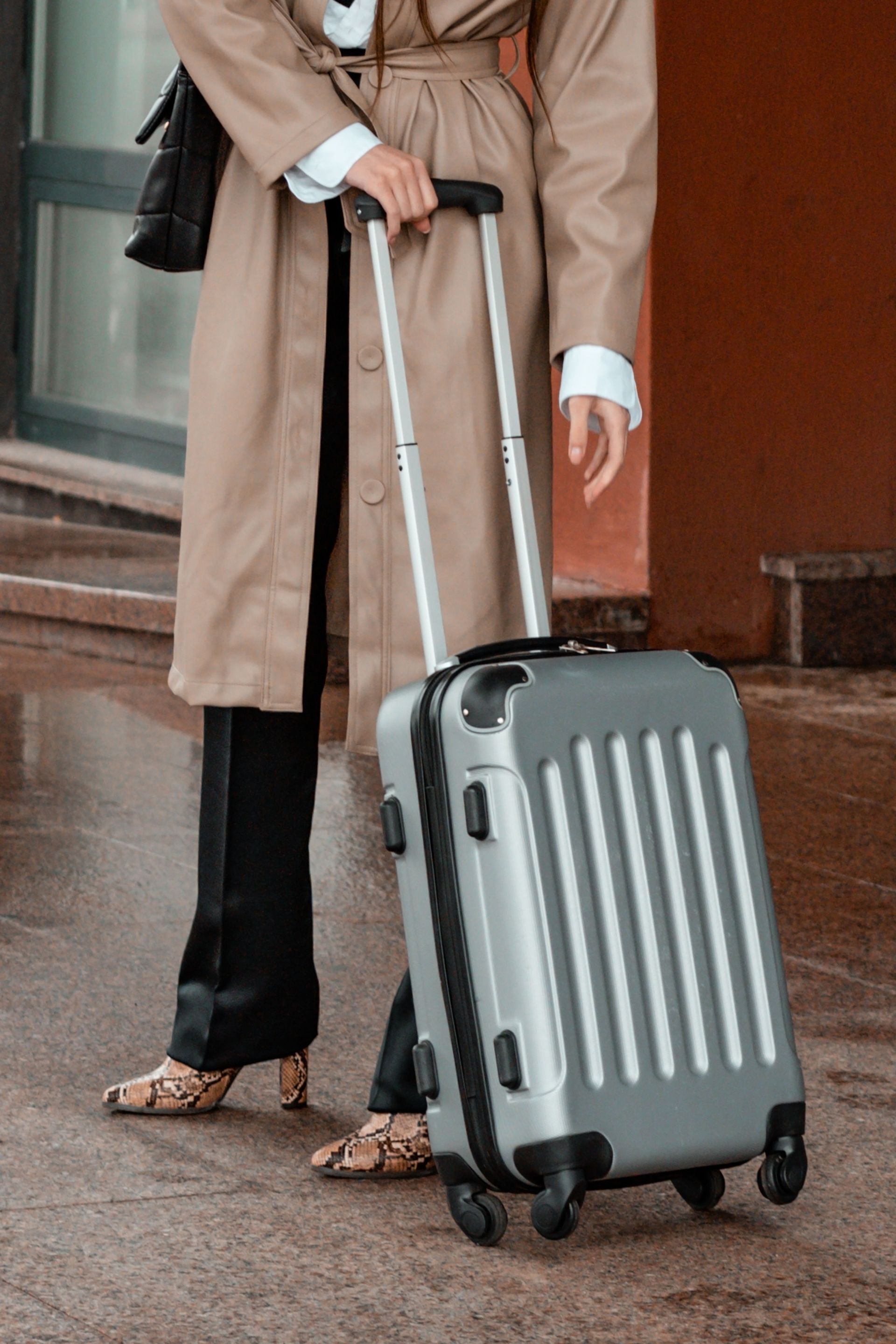 Une personne en manteau beige tire une valise grise sur un trottoir.