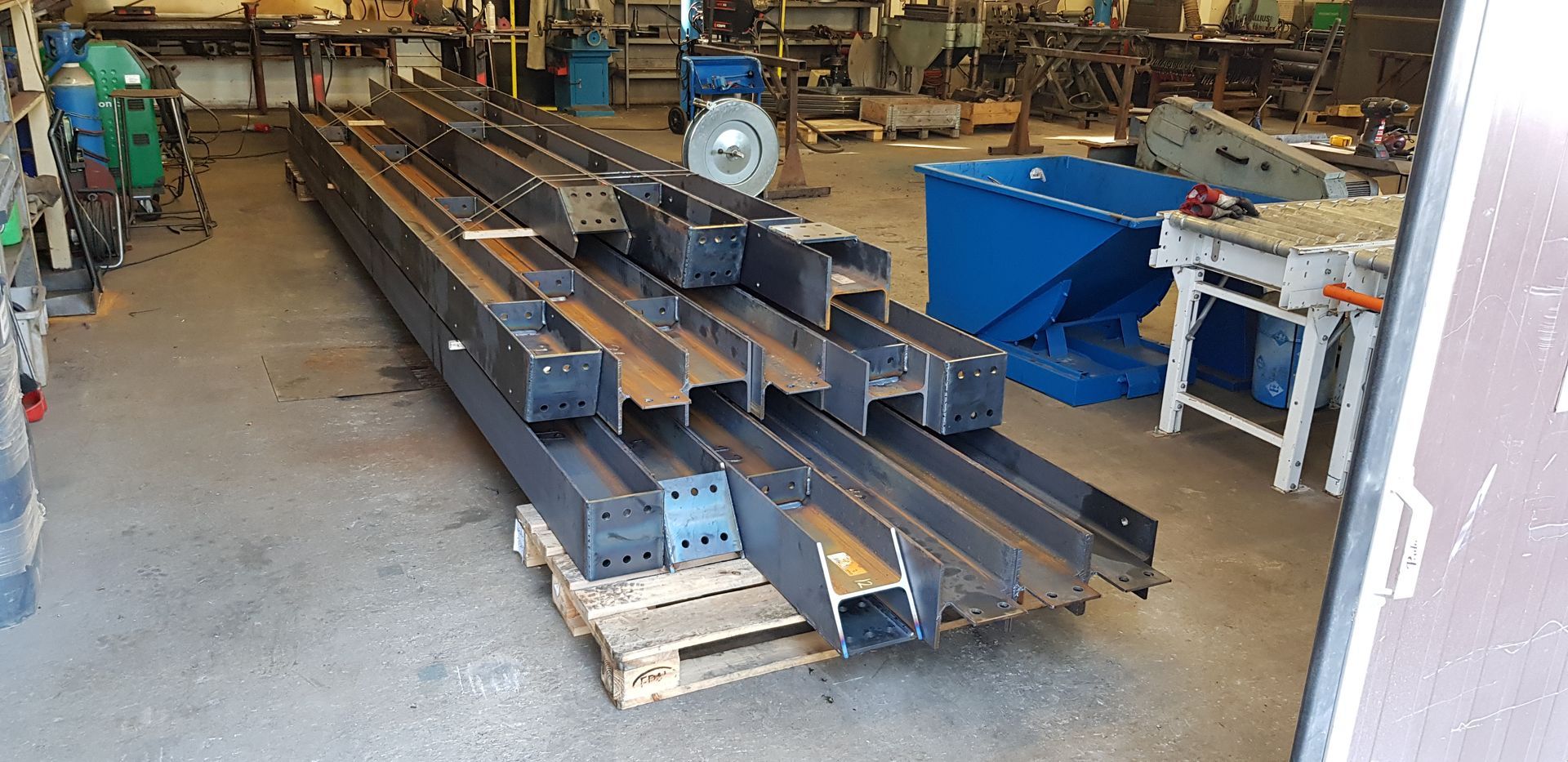Pitkiä harmaita teräspalkkeja pinottuna puisille lavoille metallityöpajassa.