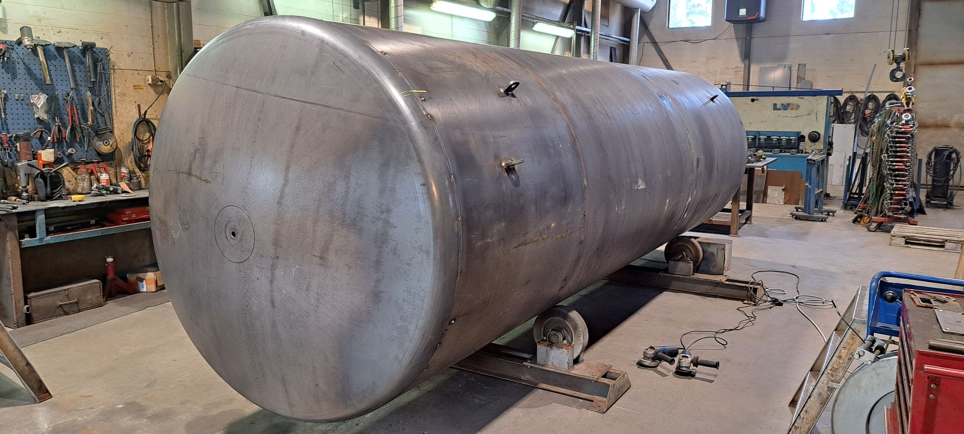 Suuri, vaakasuora, lieriömäinen metallinen varastosäiliö seisoo tukijalustoilla työpajassa, joka on täynnä työkaluja ja koneita.