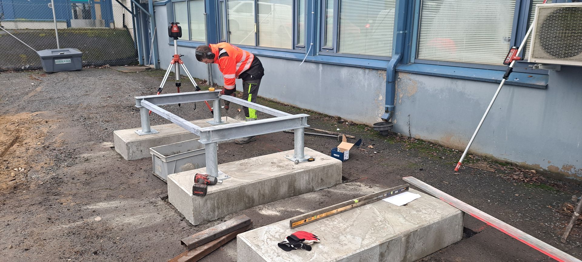 Heijastinvarusteisiin pukeutunut työntekijä säätää betonilohkoille asennettua metallirunkoa ulkona rakennustyömaalla.