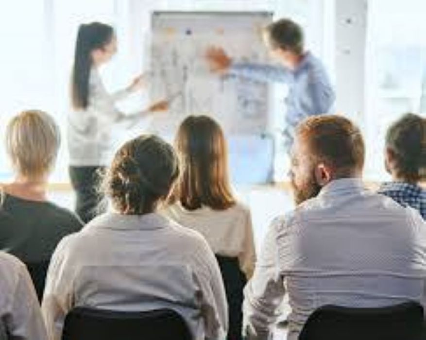 Tres personas están reunidas. Dos hombres de traje y una mujer vestida de rojo están escribiendo en una mesa.