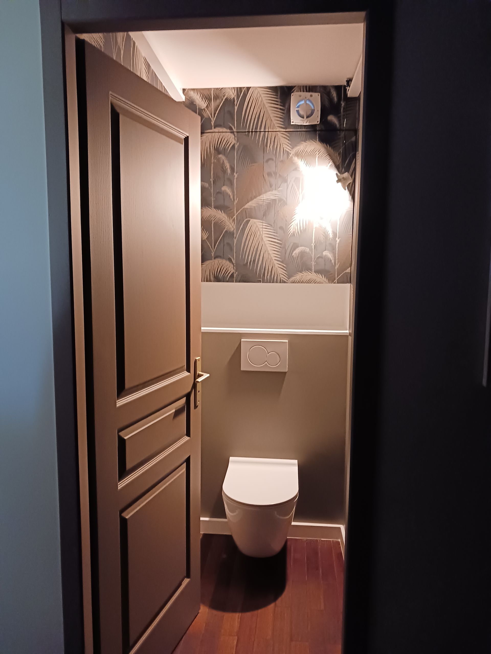 Une porte peinte en marron s'ouvre sur de petites toilettes élégantes, avec du papier peint à motifs et des toilettes blanches.