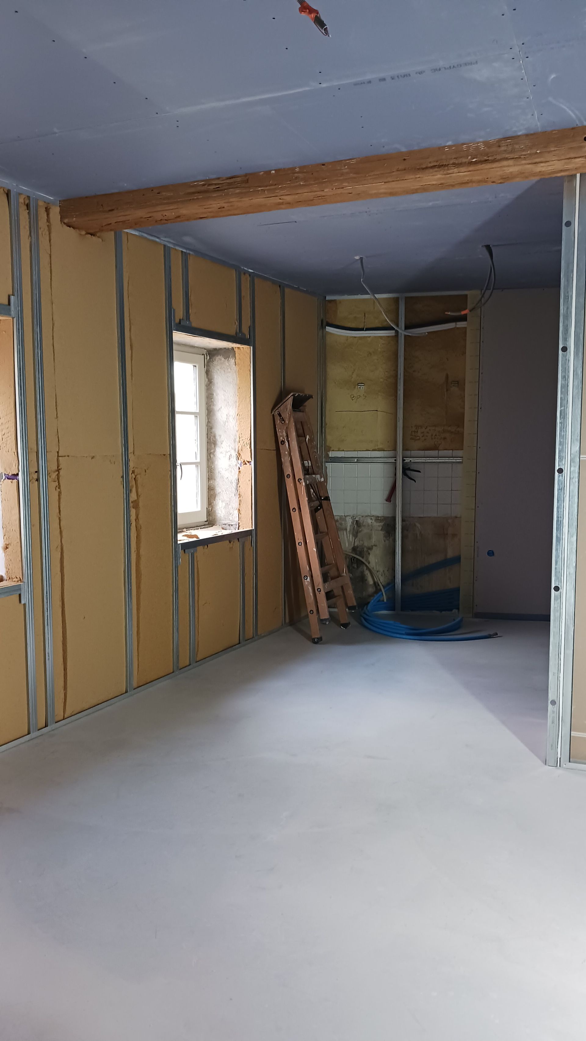 Pièce en rénovation, cloisons sèches et ossature visibles ; fenêtre avec encadrement en pierre ancienne, poutre en bois au-dessus.