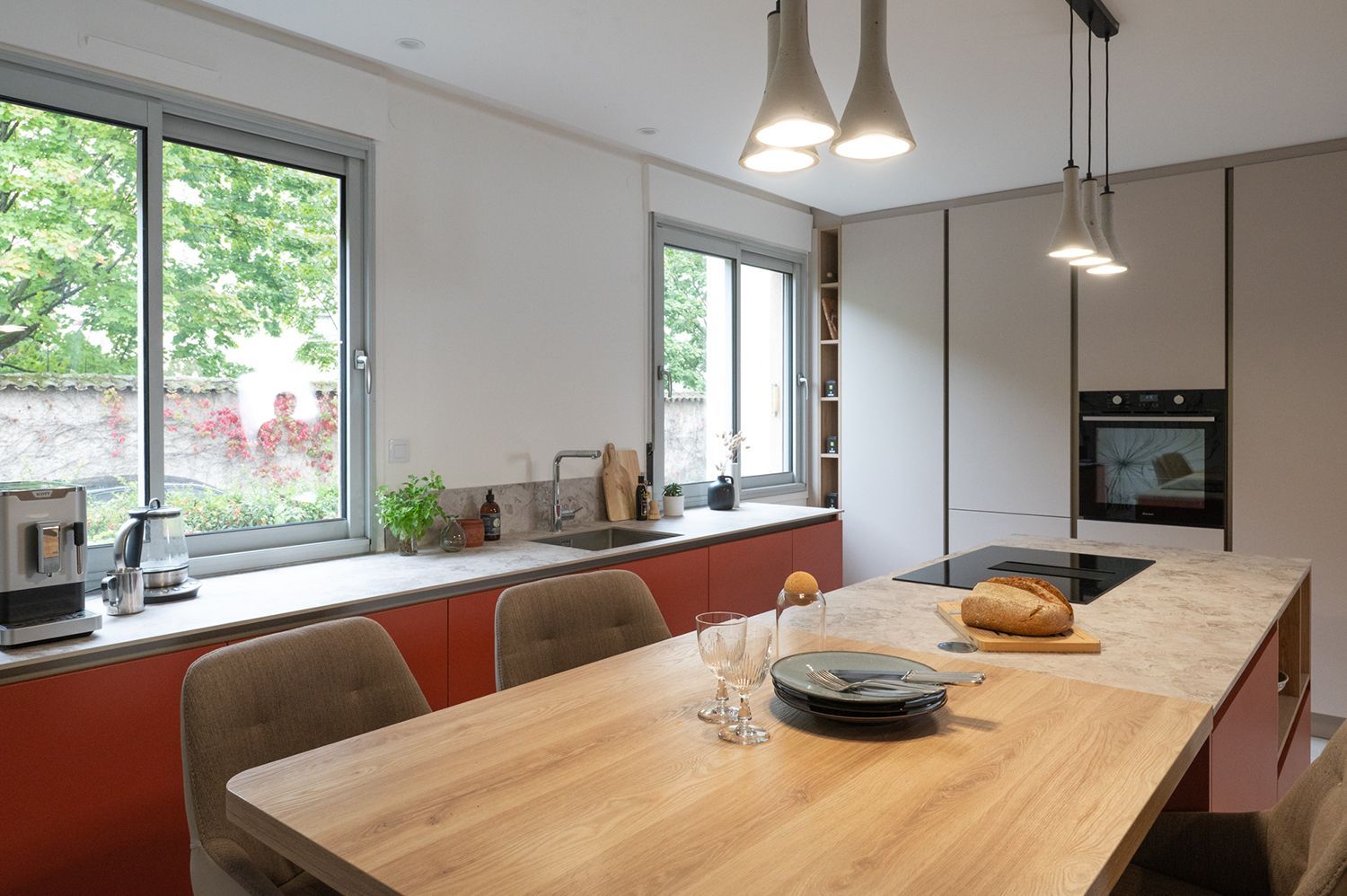 Cuisine moderne avec placards rouges, îlot en bois, grandes fenêtres et suspensions lumineuses.