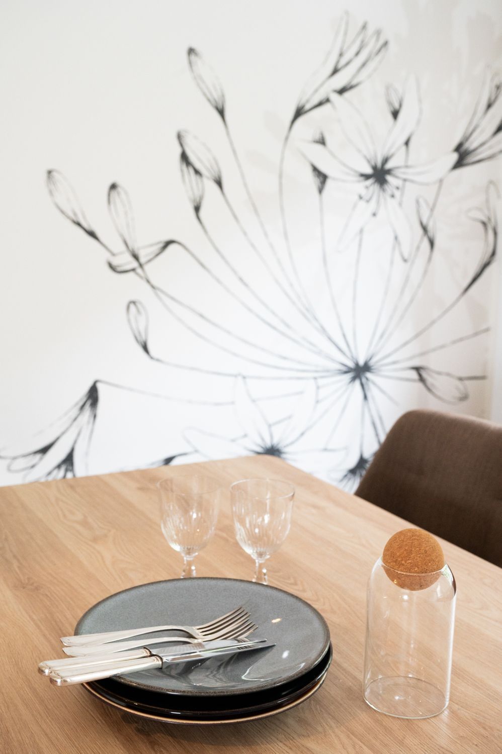 Table dressée pour le dîner avec assiettes, couverts, verres et bouteille en verre, devant une fresque murale.