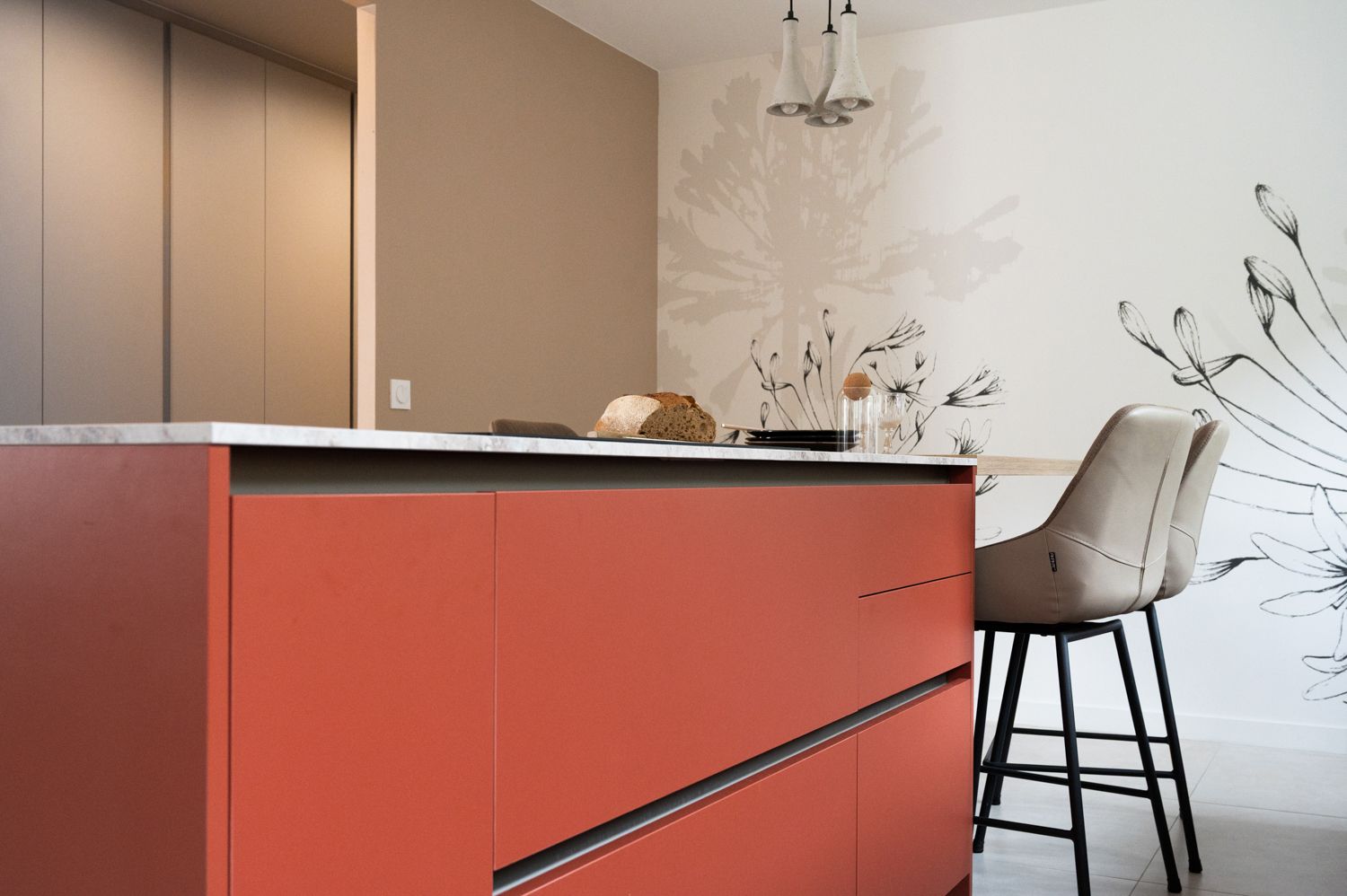 Îlot de cuisine rouge avec plan de travail blanc, murs beiges et fresque florale. Un tabouret de bar beige est placé à proximité.