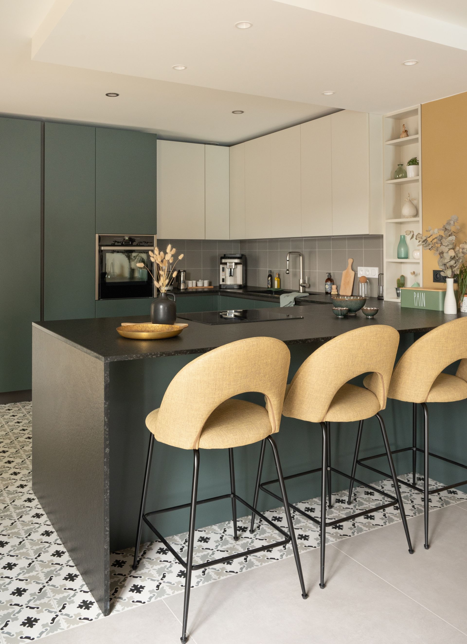 Cuisine moderne avec placards bleu sarcelle, îlot central noir avec tabourets beiges et sol carrelé à motifs.