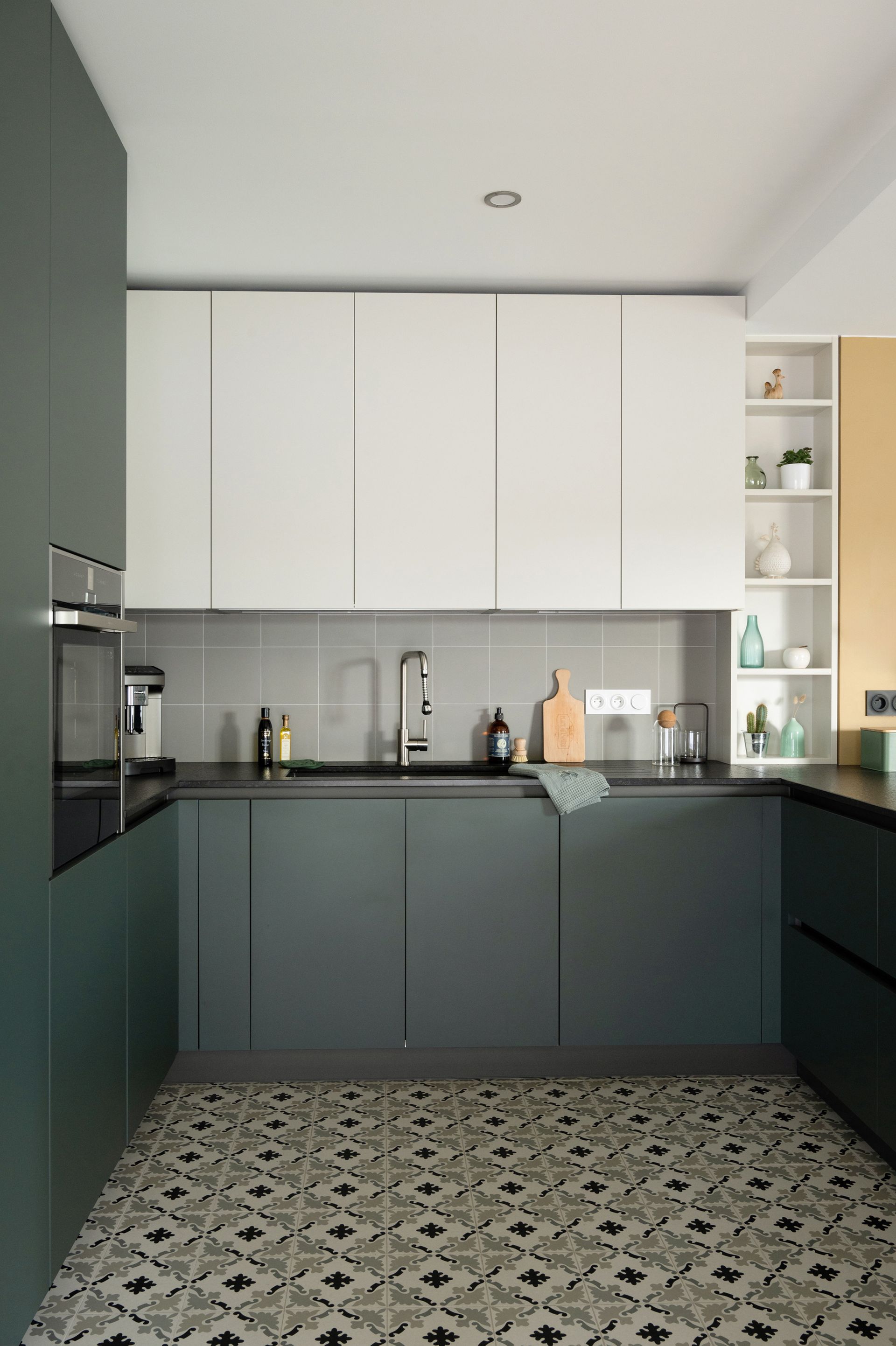 Cuisine moderne avec placards verts et blancs, crédence grise et sol carrelé à motifs.