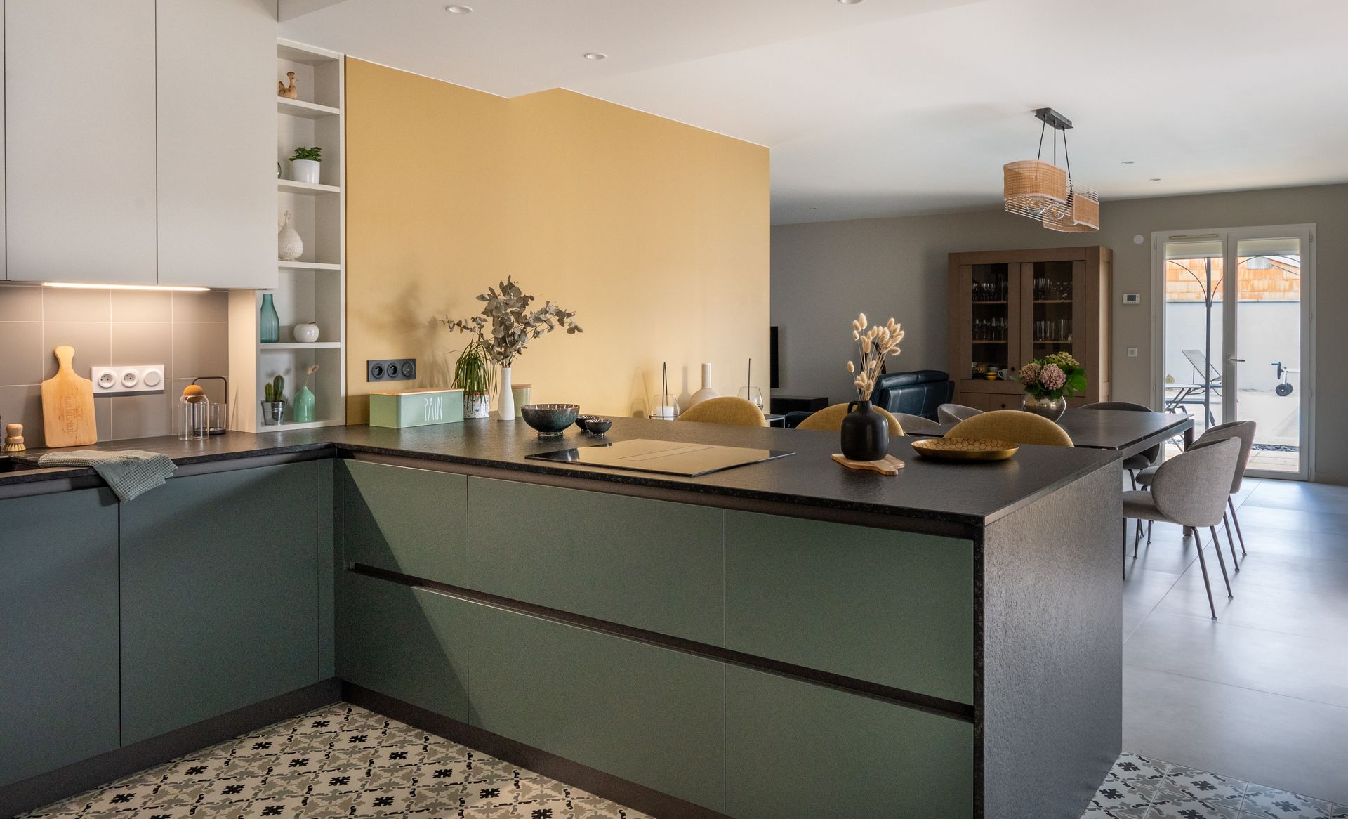 Cuisine moderne avec placards verts, plan de travail noir, ouverte sur un coin repas avec table et chaises.