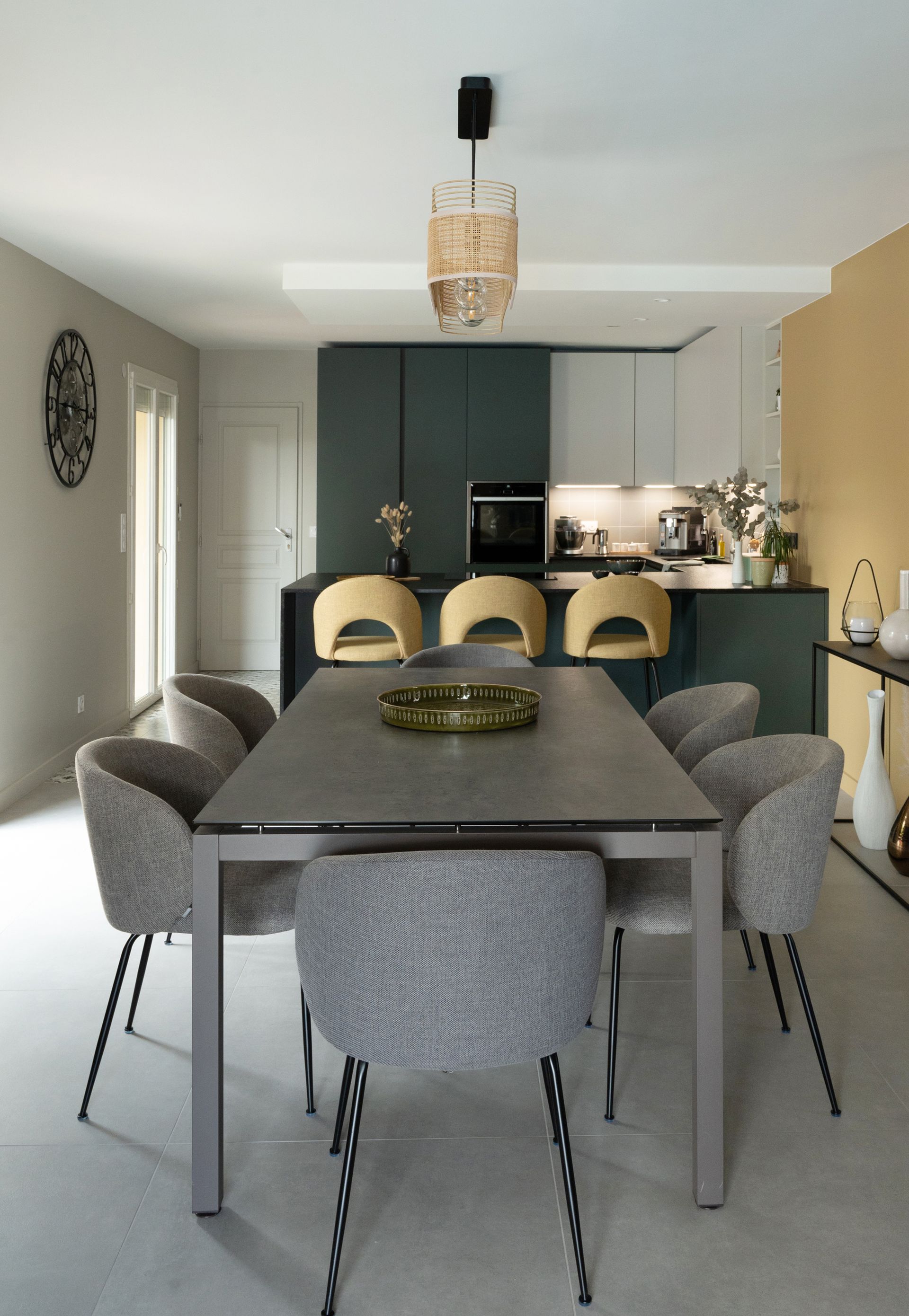 Salle à manger avec table et chaises grises, cuisine en arrière-plan. Placards bleu sarcelle, mur jaune et tabourets beiges.